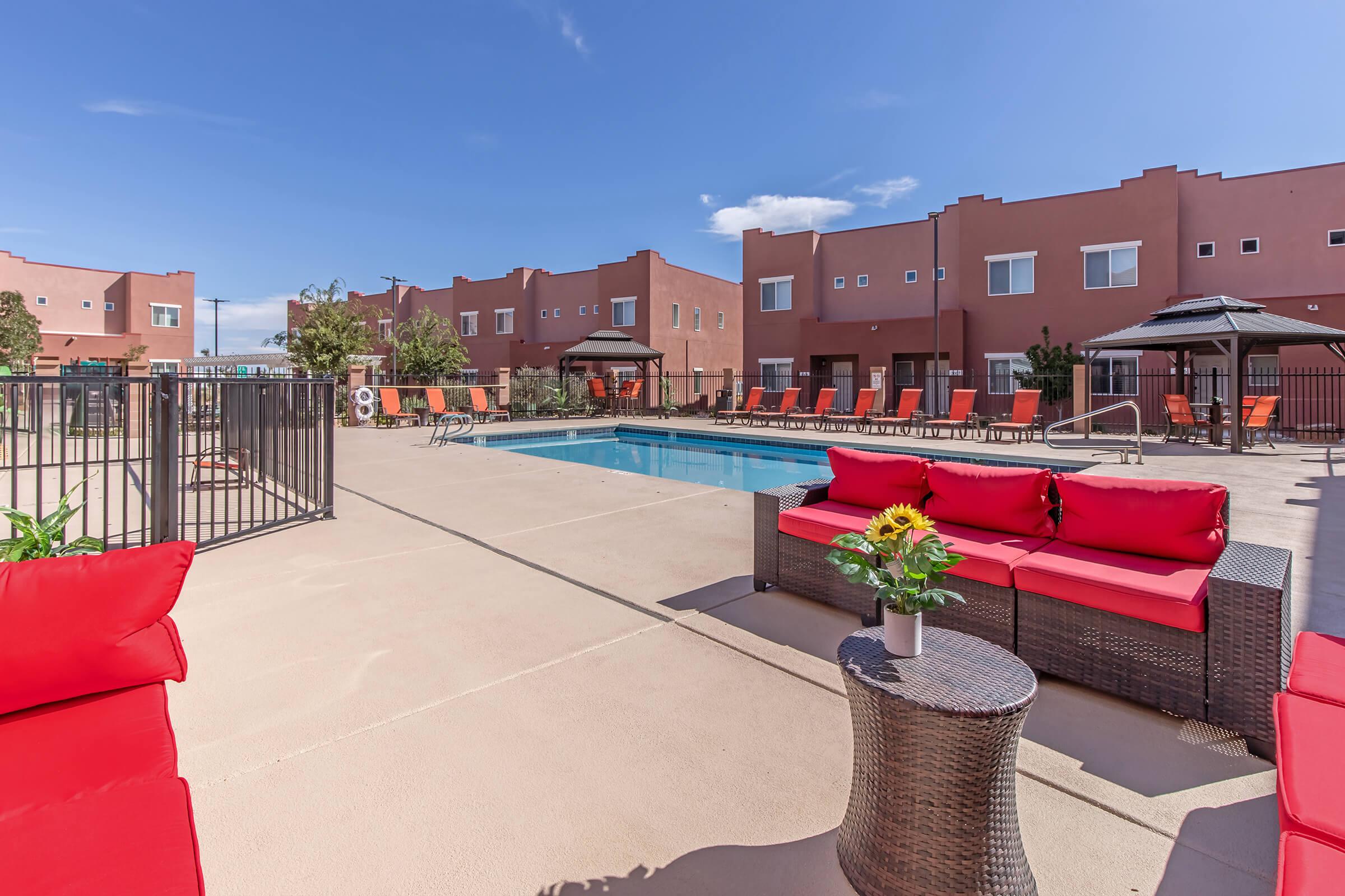 A sunny pool area featuring a swimming pool surrounded by lounge chairs. In the foreground, bright red sofas and a small table with a vase of sunflowers create a welcoming atmosphere. The backdrop consists of light-colored buildings under a clear blue sky, adding to the relaxing ambiance.