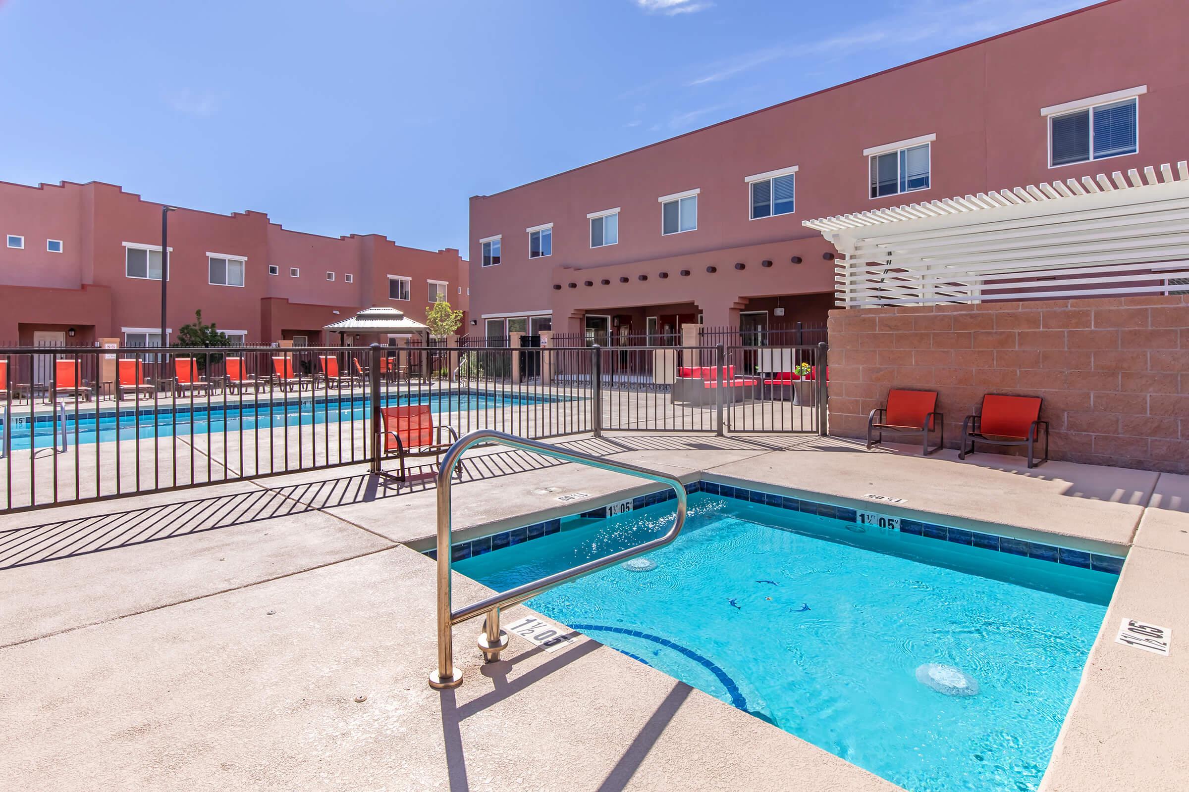 A clear blue swimming pool surrounded by a concrete deck, featuring a handrail leading down into the water. Bright orange lounge chairs are positioned around the pool area. In the background, there are two multi-story buildings, with a clear blue sky above.