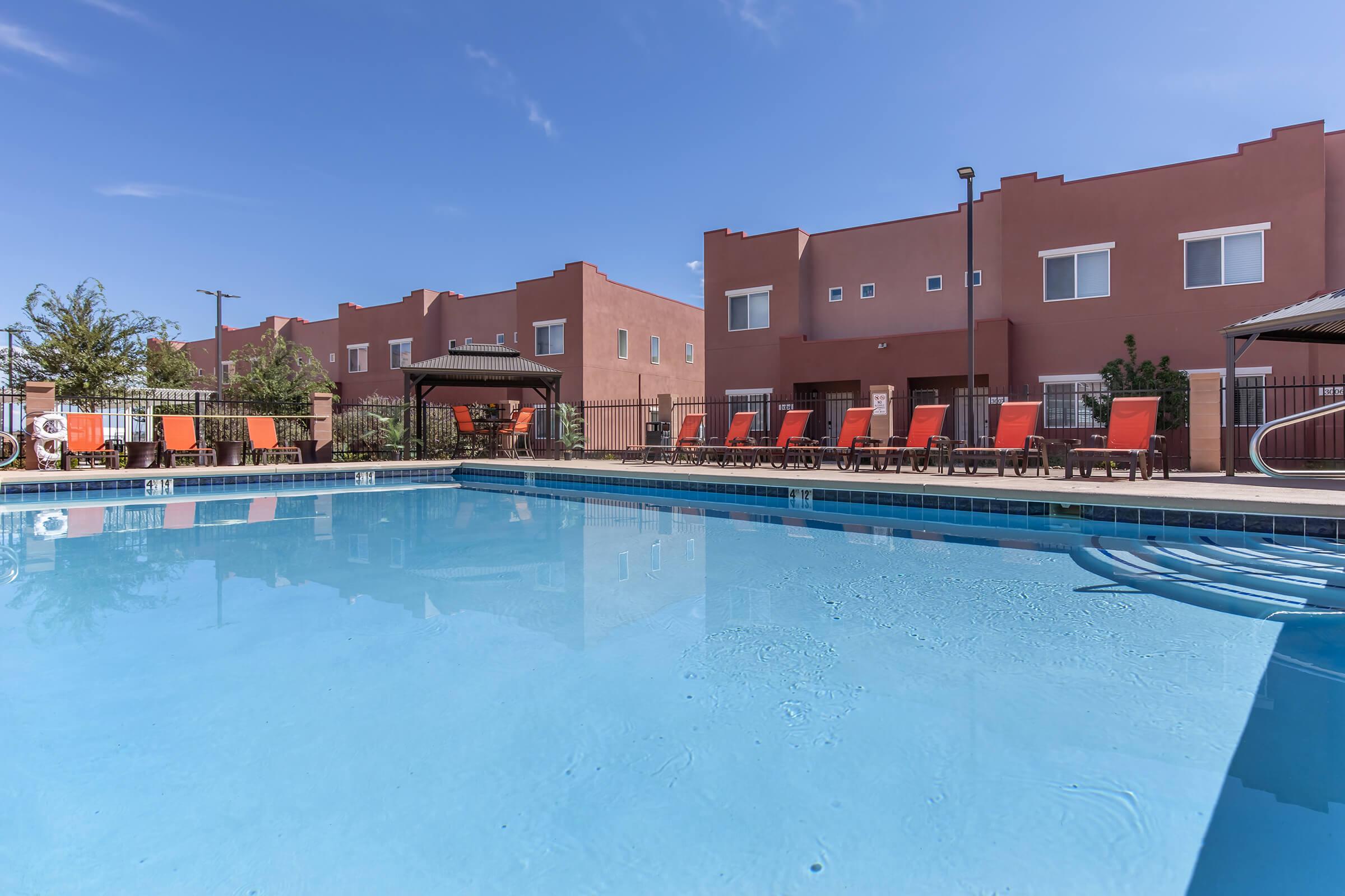 A clear blue swimming pool surrounded by orange lounge chairs. In the background, there are brown buildings with multiple windows and a small gazebo to the side. The sky is bright blue with a few clouds, and there are some trees and plants nearby, adding a touch of greenery to the scene.