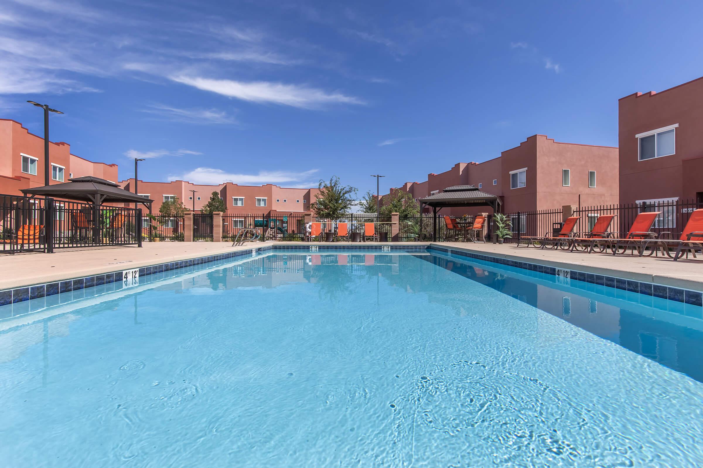 A clear blue swimming pool surrounded by lounge chairs and umbrellas, set against colorful adobe-style buildings under a bright blue sky with a few clouds. The area is inviting and well-maintained, ideal for relaxation and leisure.