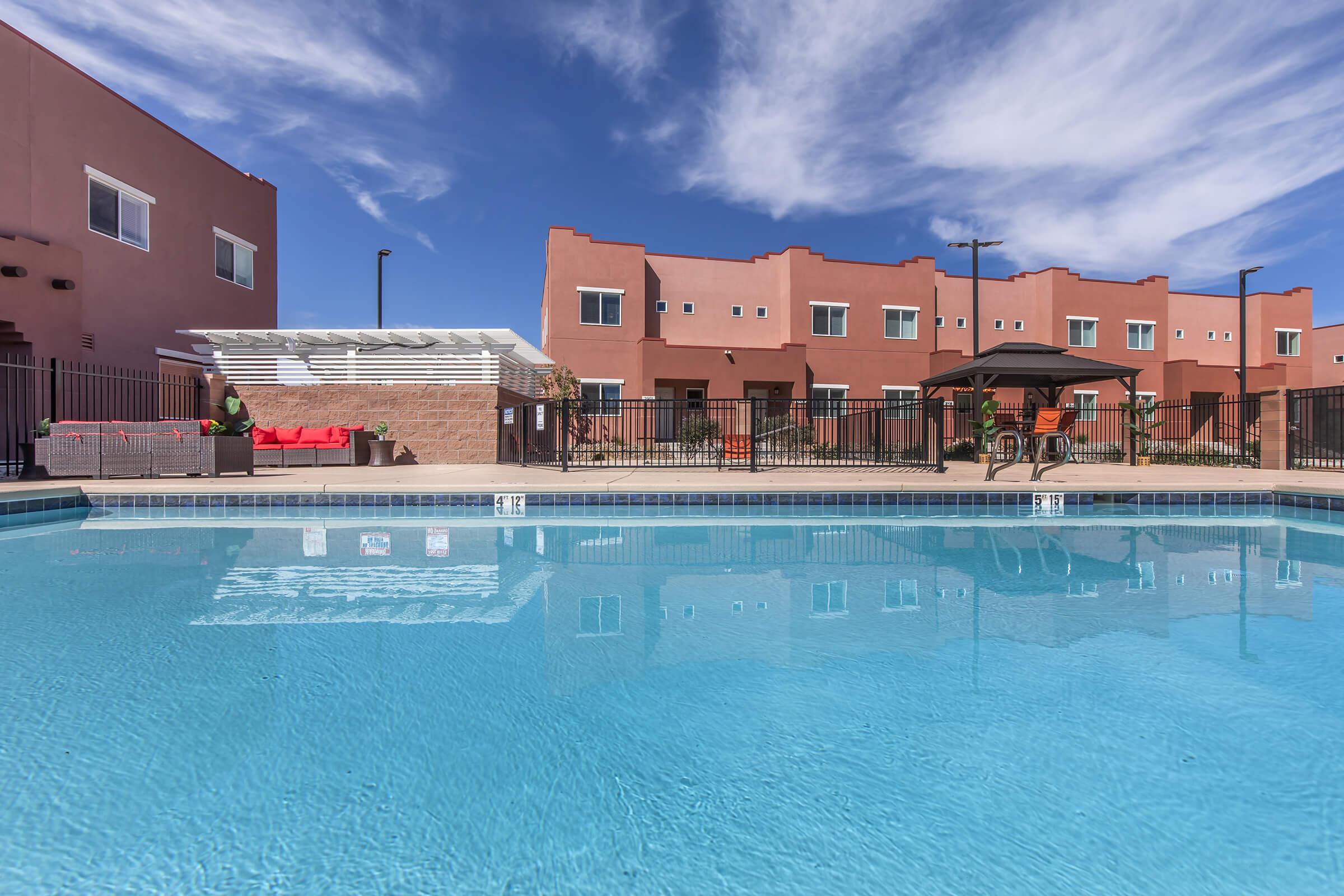 A clear blue swimming pool reflects the sky, surrounded by a fence. In the background, there are two-story buildings with a reddish exterior. A covered seating area with lounge furniture is visible near the poolside, alongside a shaded gazebo, creating a relaxed outdoor atmosphere.