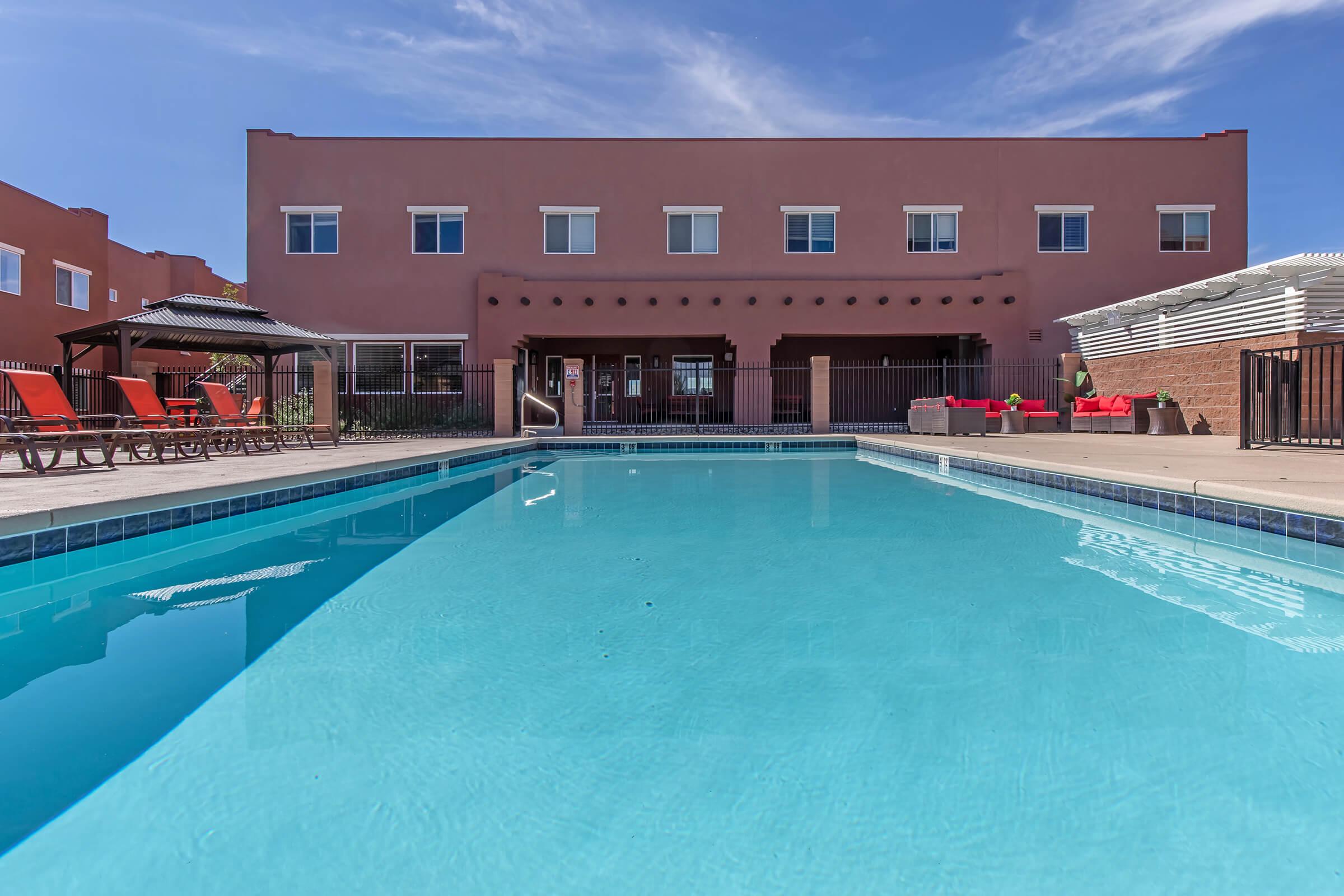A clear blue swimming pool in front of a modern building with a pink exterior. The pool area features lounge chairs and shaded seating. The sky is bright with a few clouds, creating a vibrant and inviting atmosphere for relaxation.