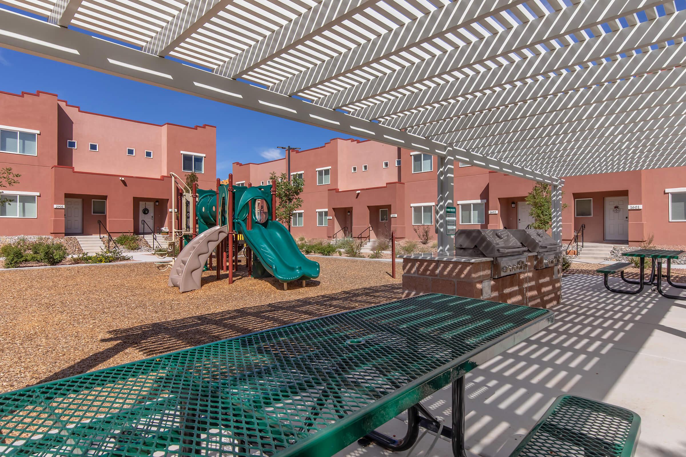 A courtyard area featuring a playground with slides, surrounded by residential buildings. There are shaded picnic tables beneath a pergola and landscaped areas with plants. The sky is clear and blue, enhancing the inviting atmosphere of the space.