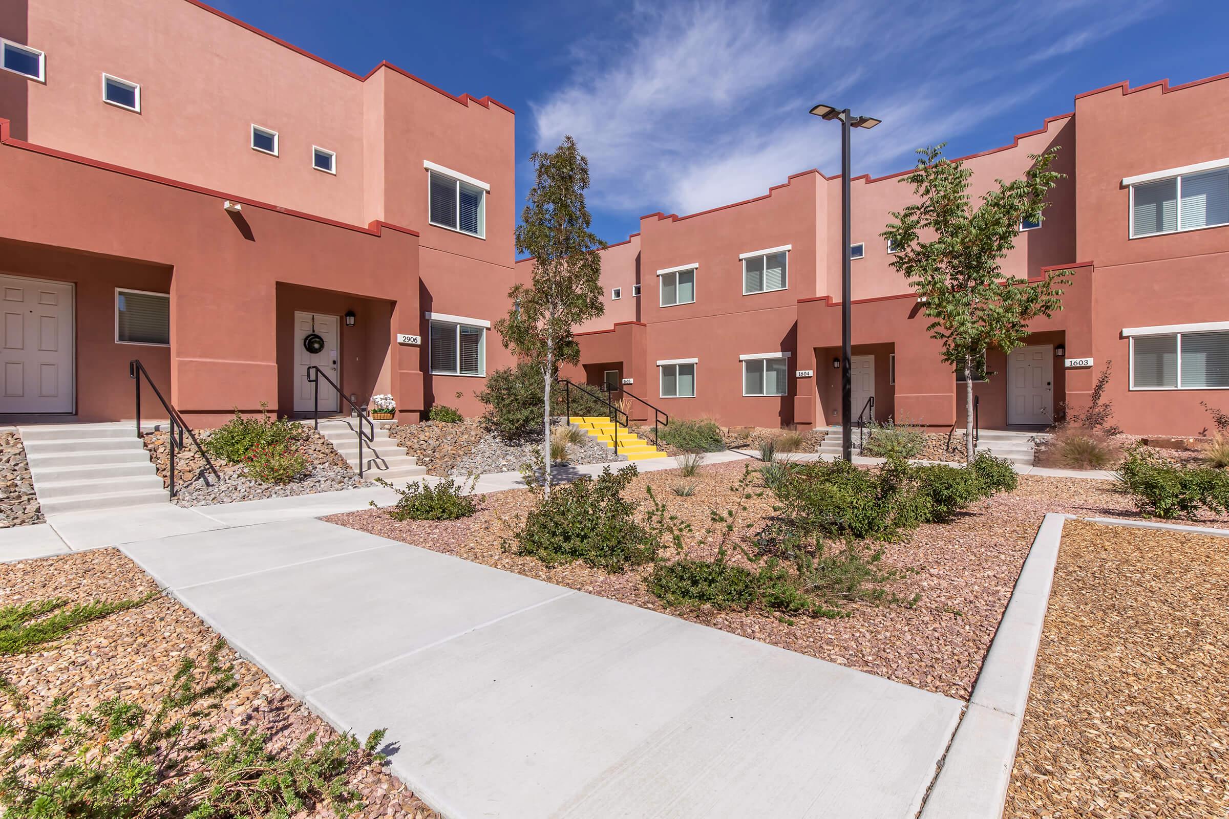 A modern residential complex featuring several connected buildings with a warm color palette. Each unit has a small front porch, and there are landscaping elements like trees and shrubs. Pathways made of concrete lead to the entrances, set against a clear blue sky.