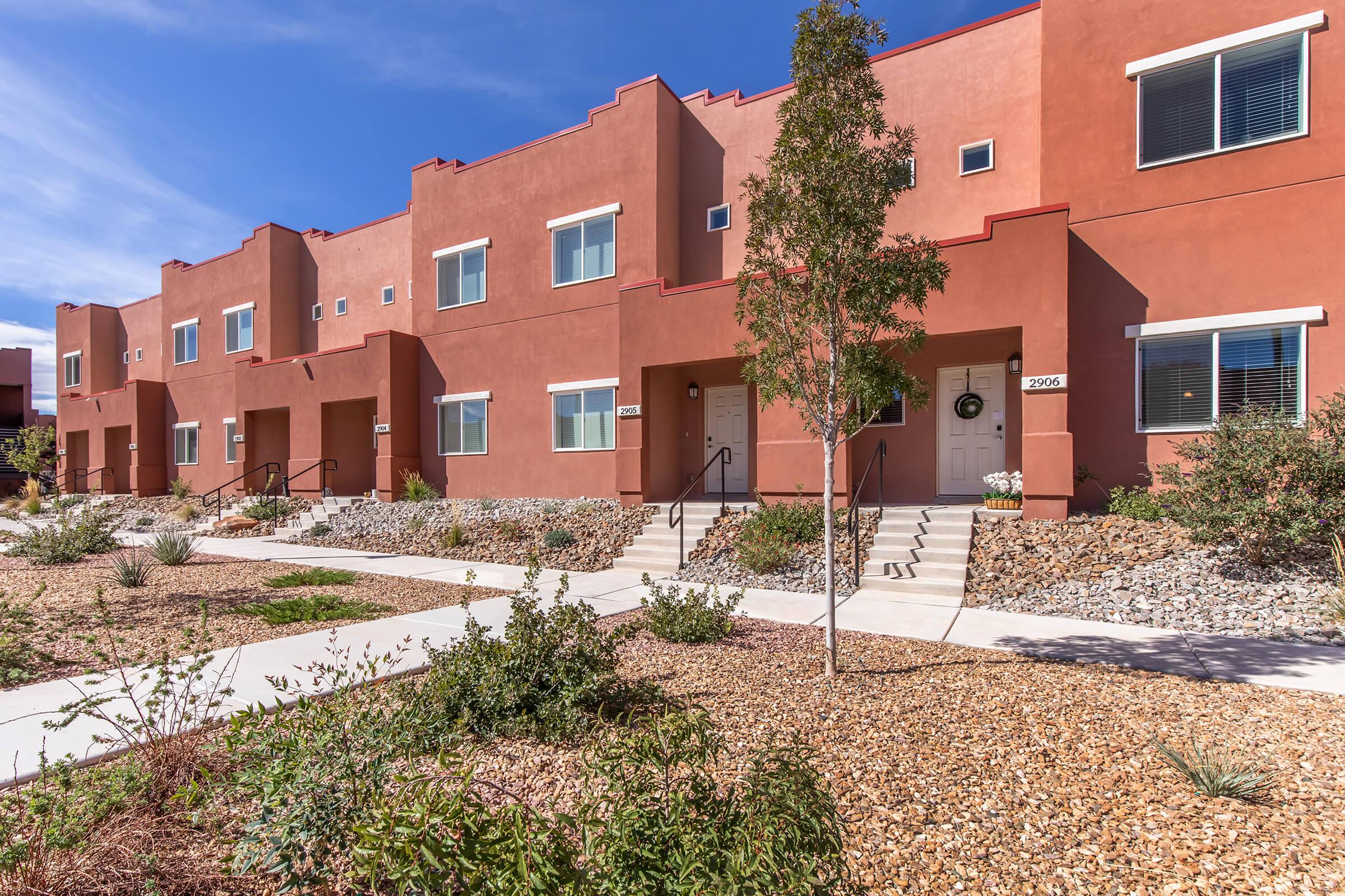 A row of modern, two-story townhomes with red stucco exteriors and white trim. Each unit has a small porch with a staircase leading up to the front door. The landscaping includes drought-resistant plants and rocks, with the sky visible in the background.