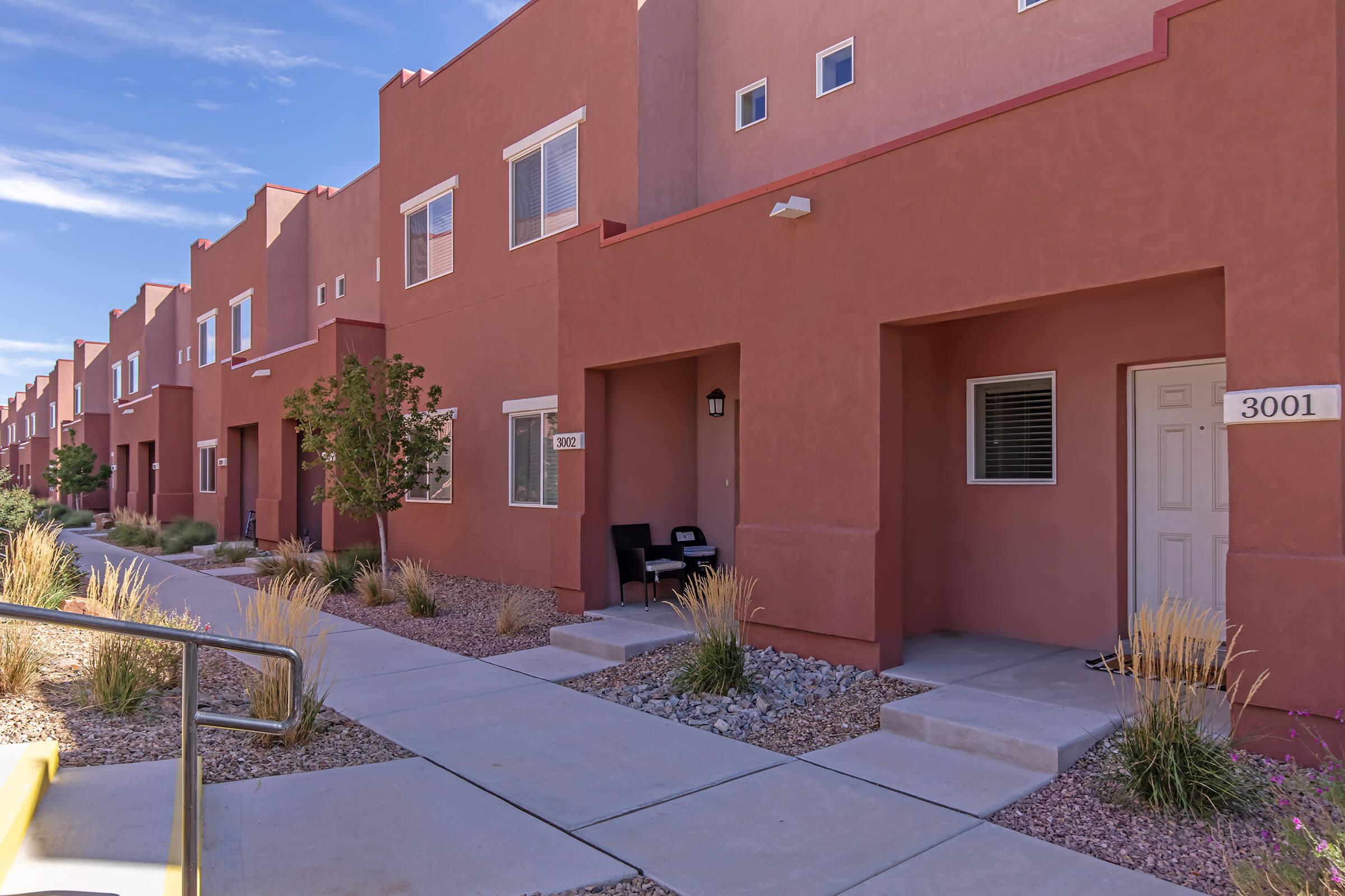 Row of modern, two-story townhouses with earthy red exteriors and simple architectural design. Each unit features a small porch with seating and landscaped areas filled with low-maintenance plants. Clear blue sky above, creating a bright and inviting atmosphere.