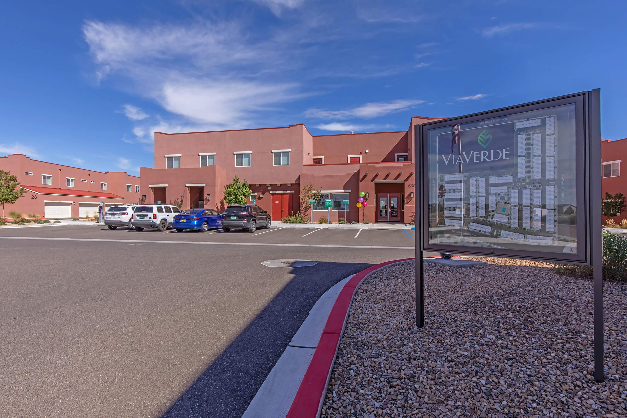 A modern building with a reddish exterior and multiple windows, featuring a large sign that reads "VIA VERDE." The foreground includes a parking lot with several cars, and the scene is set under a clear blue sky, with decorative landscaping along the edges.