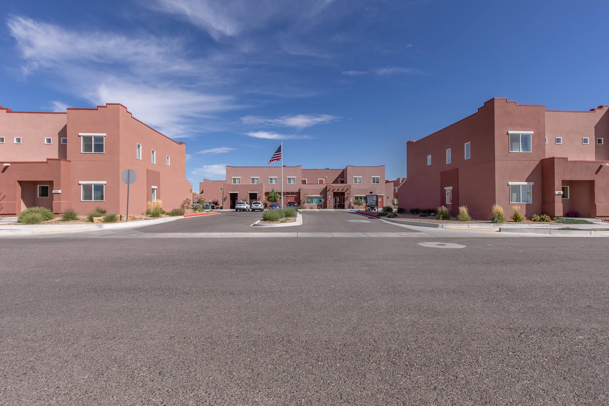 View of a residential area featuring adobe-style buildings painted in warm tones. The scene includes a clear blue sky with scattered clouds, an American flag flying prominently, and vehicles parked in the courtyard area. The layout suggests a peaceful neighborhood atmosphere.