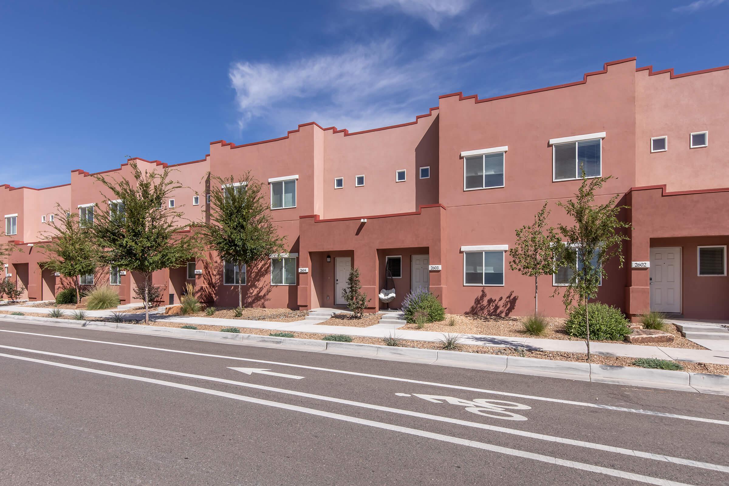A row of modern, adobe-style townhouses with light brown walls and landscaped small gardens. Each unit features windows, a front door, and trees planted along the walkway. The street in front includes bicycle markings and is clear, under a blue sky with some clouds.