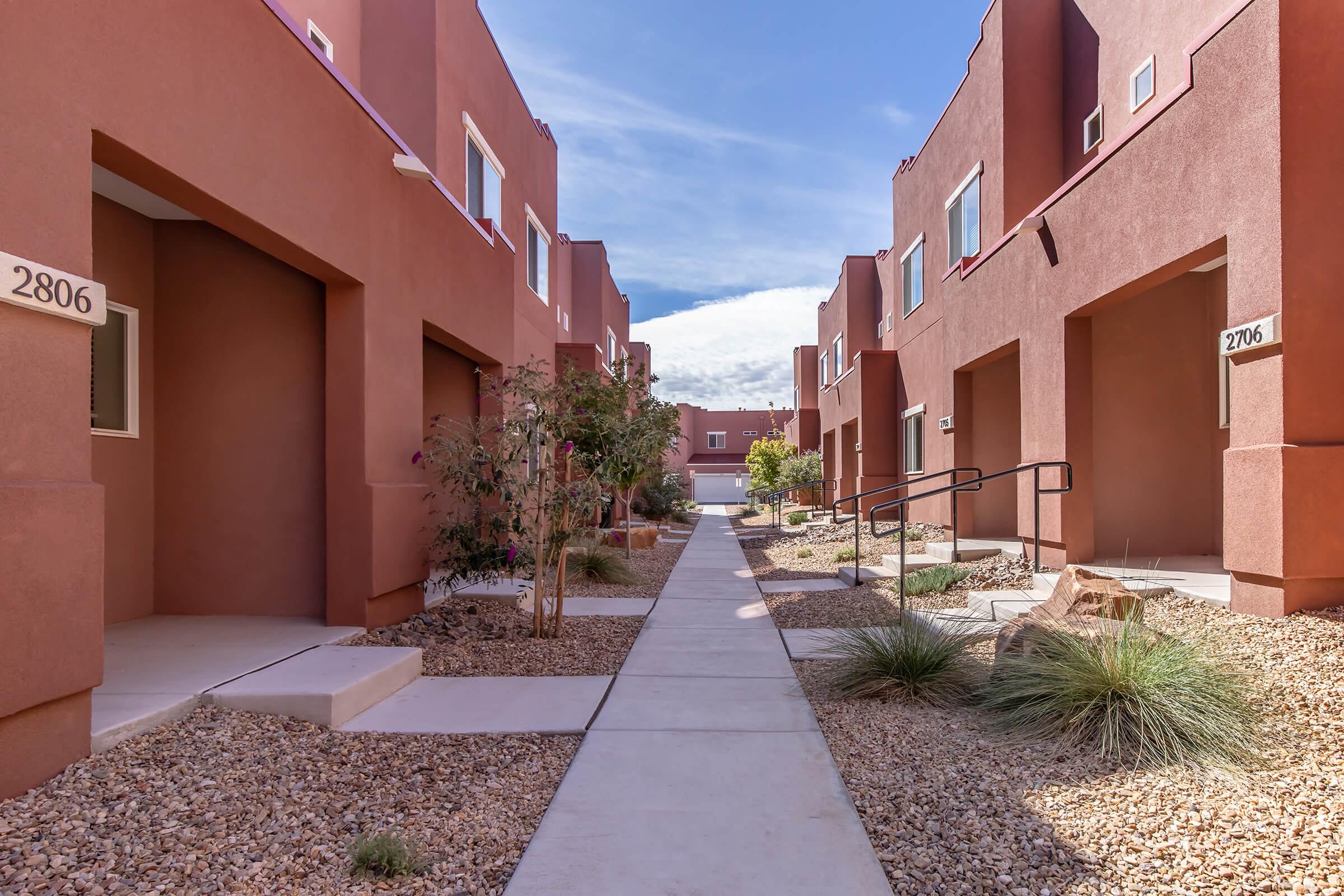 A walkway between modern, reddish-brown residential buildings. There are small shrubs and gravel landscaping along the path, with doorways visible on either side. The sky is clear with scattered clouds.