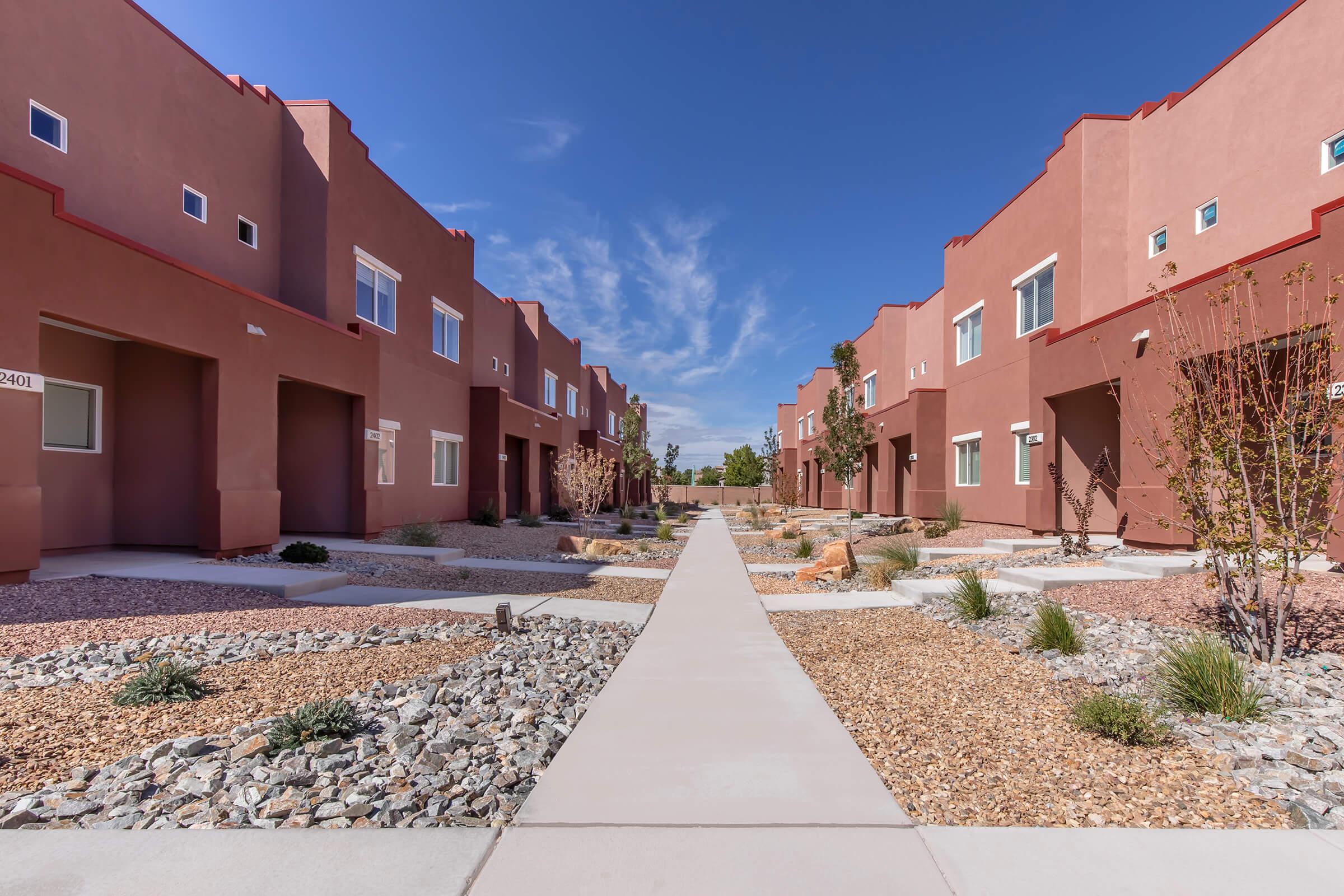 A modern residential street lined with reddish-brown townhouse units, featuring a straight concrete pathway. The area has desert landscaping with gravel, small shrubs, and trees under a clear blue sky with wispy clouds.