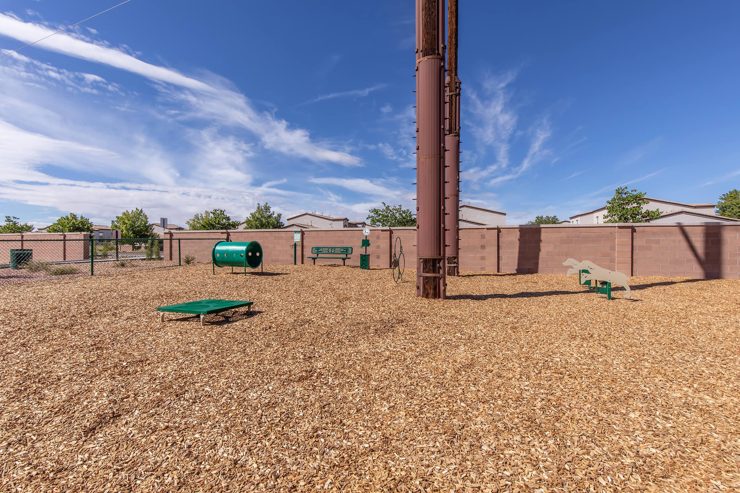A spacious dog park with wooden chip ground, featuring several green agility stations, including a tunnel and elevated platforms, surrounded by a low brick wall under a clear blue sky with a few clouds.