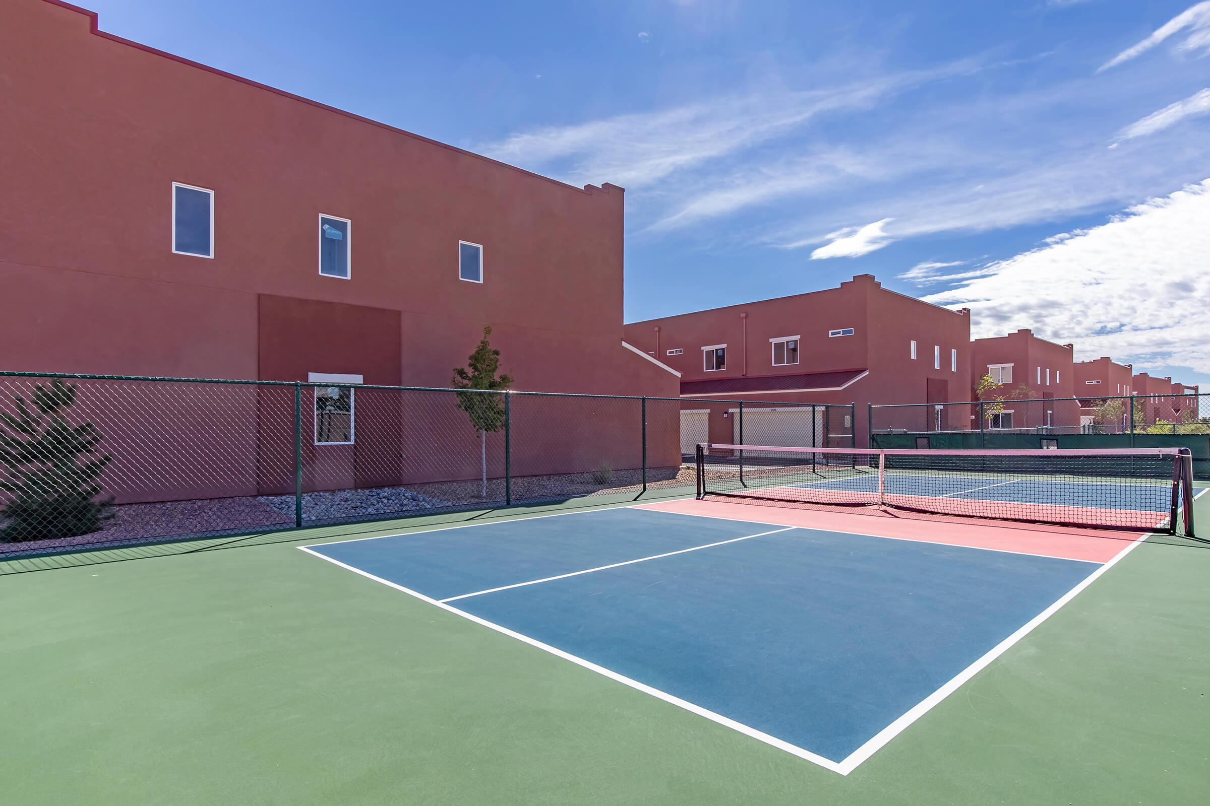 A view of a tennis court with a green playing surface and a red court in the foreground. Behind the courts are buildings painted in earthy tones under a clear blue sky with scattered clouds. The area is enclosed with a black fence, and there are a few trees near the courts.