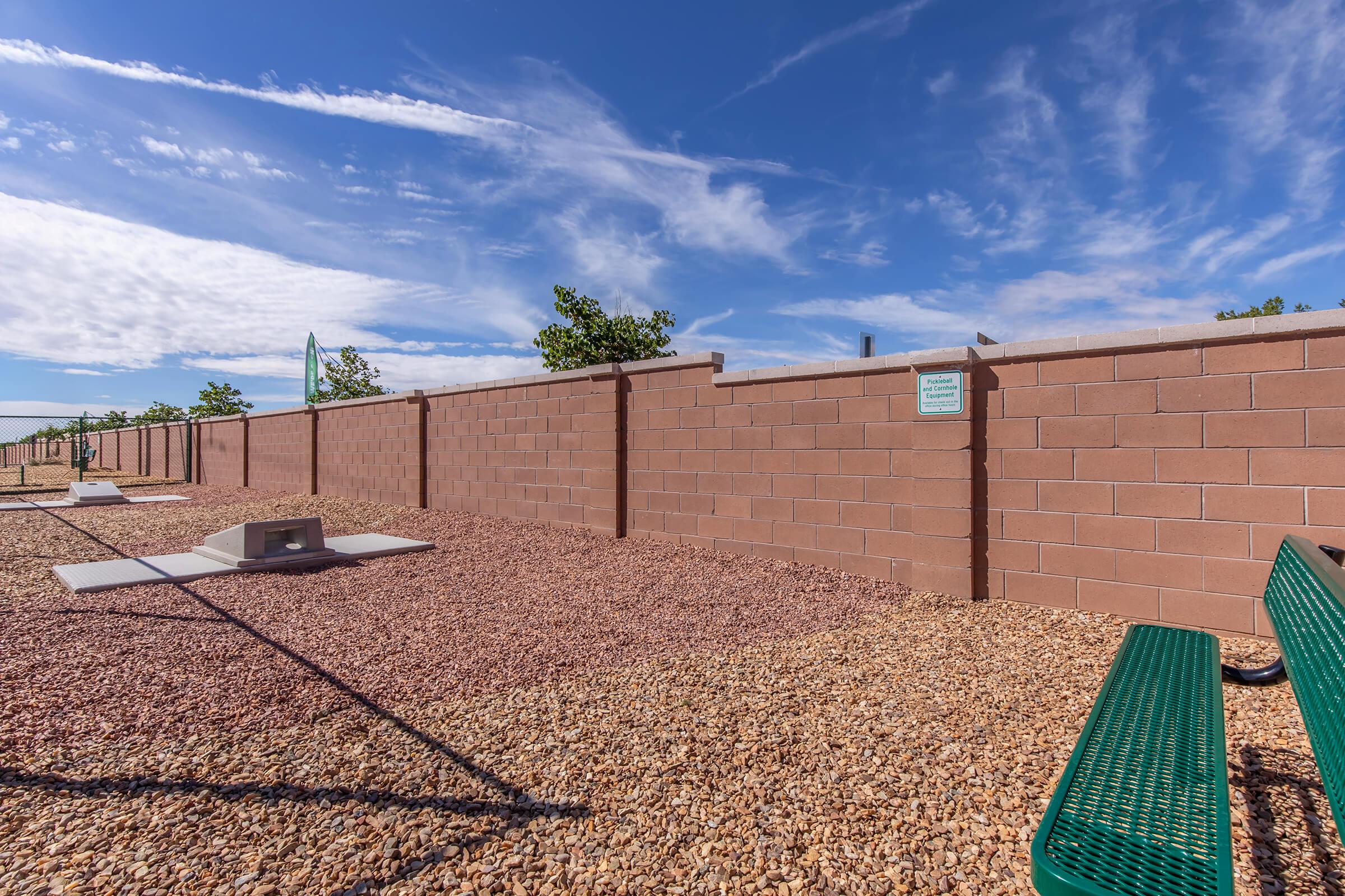 A clear blue sky with wispy clouds above a low, tan brick wall. In the foreground, a gravel-covered area features green metal benches and a concrete slab, suggesting a public park or recreational space. The scene is bright and sunny, evoking a tranquil outdoor environment.