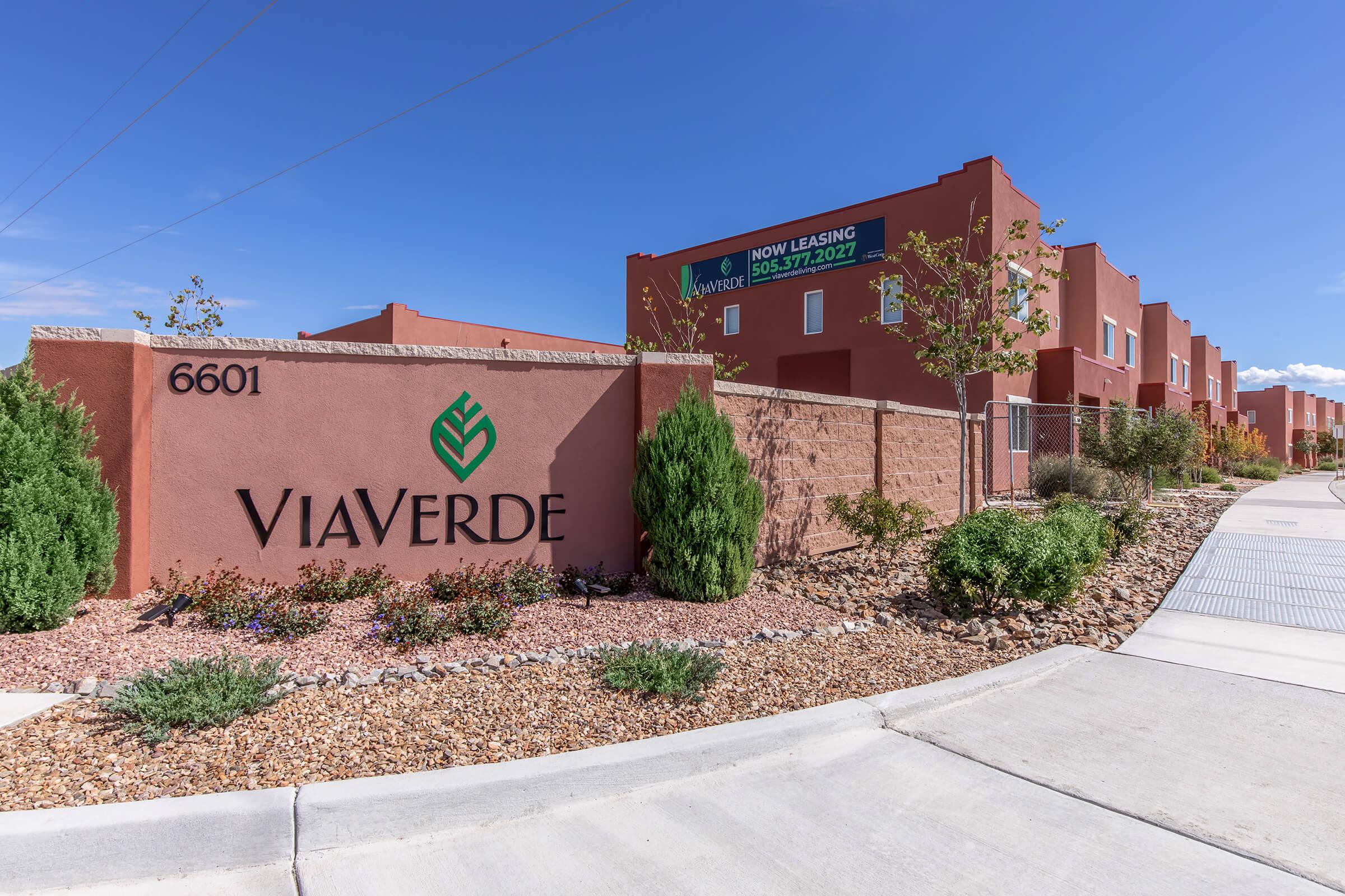 A stone sign displaying "VIAVERDE" in front of a modern apartment complex, with the address "6601" visible. The background features several colorful buildings and a clear blue sky. There are well-maintained walkways, shrubs, and decorative landscaping around the sign. A banner indicates that apartments are available for leasing.