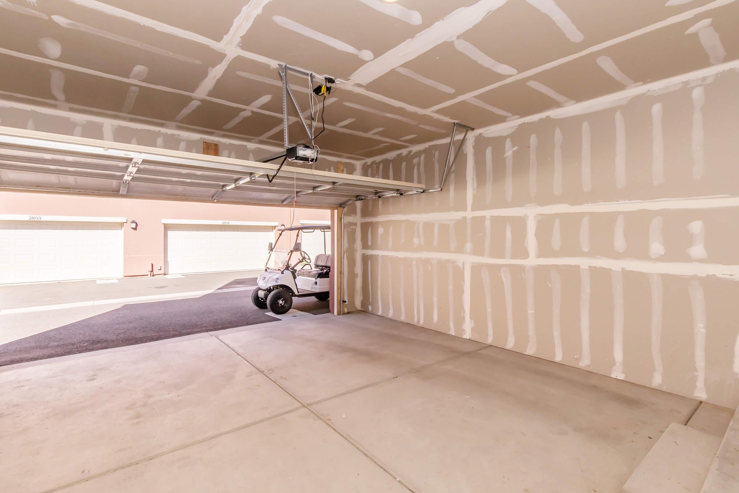 Empty garage interior with drywall that has not been finished, featuring exposed seams and a concrete floor. A white golf cart is parked in the corner near the garage door, which is open to the outside. The walls show signs of being prepared for painting or finishing, while the floor is plain and unobstructed.