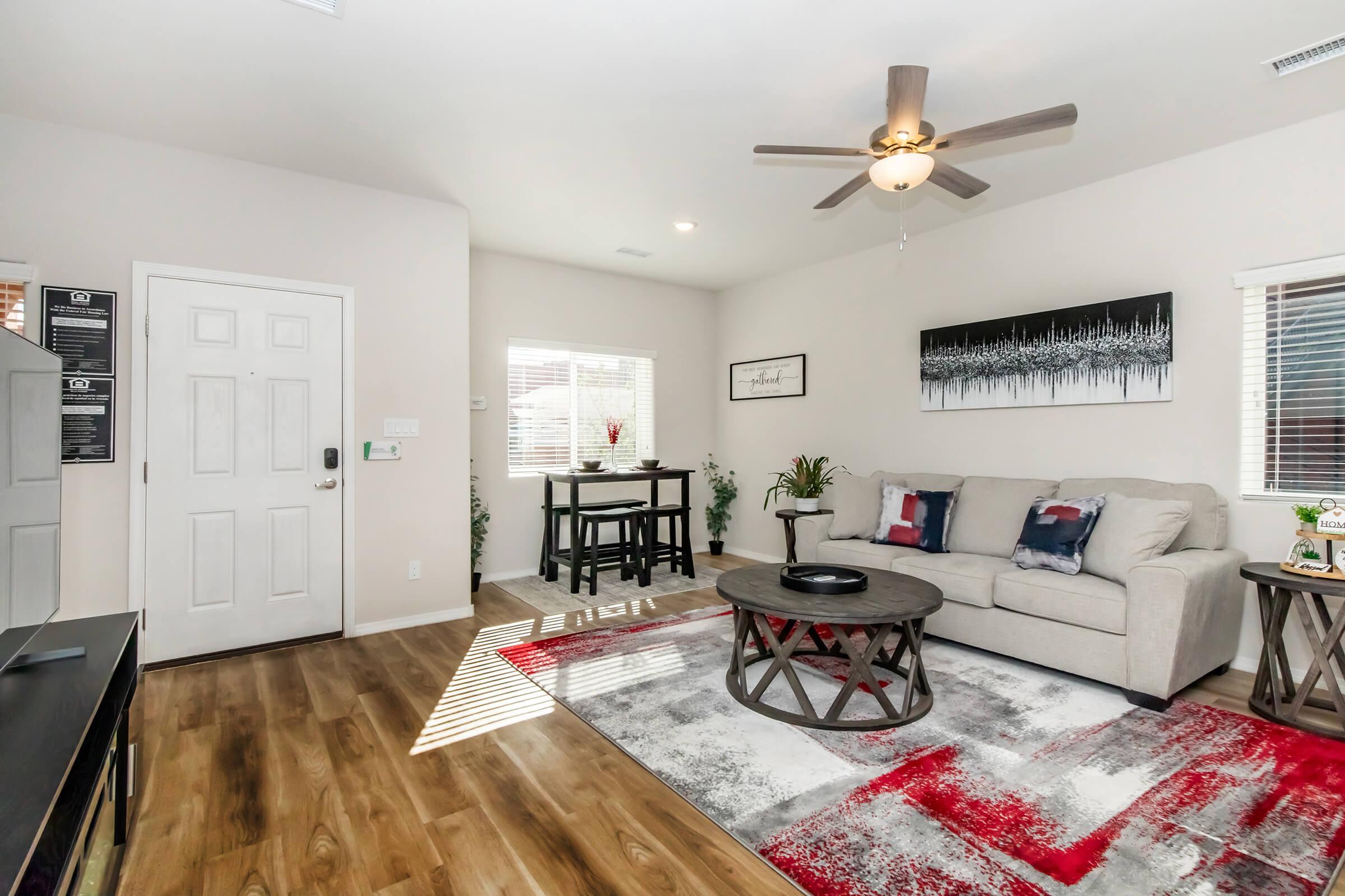 A cozy living room featuring light-colored walls, a beige sofa with decorative pillows, a round coffee table on a patterned rug, and a small dining area with a two-person table. Sunlight streams through windows, with plants and wall art adding a touch of warmth and style.