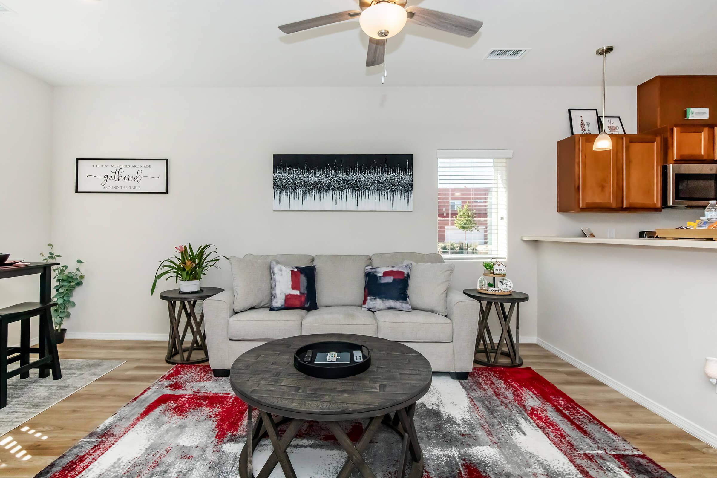 A modern living room featuring a light gray sofa with colorful throw pillows, a round coffee table, and a patterned rug. A fan is overhead, and a painting hangs on the wall. Natural light comes through a window, while plants and decorative items add a personal touch to the space.