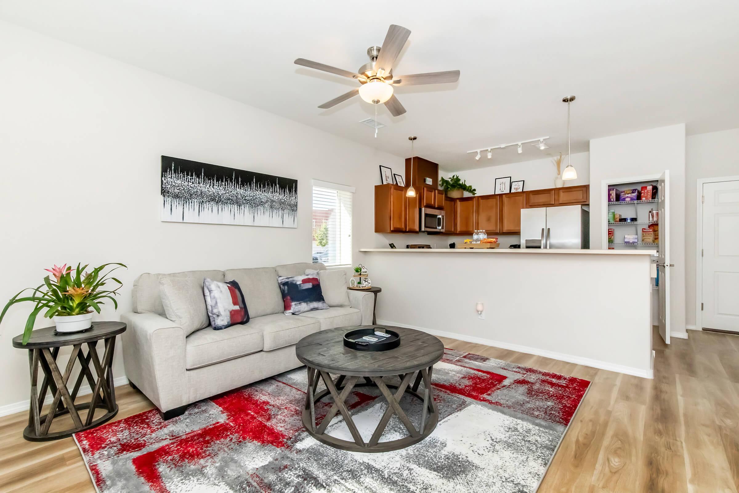 Cozy living room featuring a light gray sofa with colorful throw pillows, a round coffee table on a patterned rug, and a ceiling fan. The open kitchen in the background has wooden cabinets and modern appliances, with natural light streaming in through a window. Decor elements include a plant and wall art.