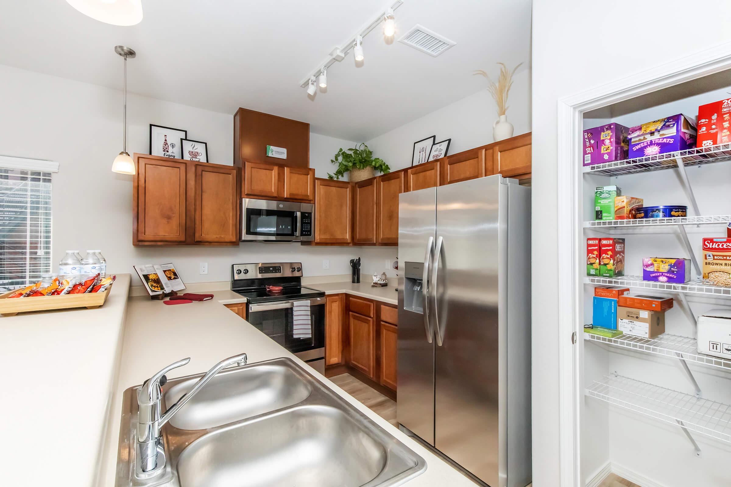 A modern kitchen featuring wooden cabinets, stainless steel appliances including a microwave and refrigerator, a double sink, and a pantry stocked with various food items. Natural light floods the space through a window, and decorative plants enhance the ambiance.