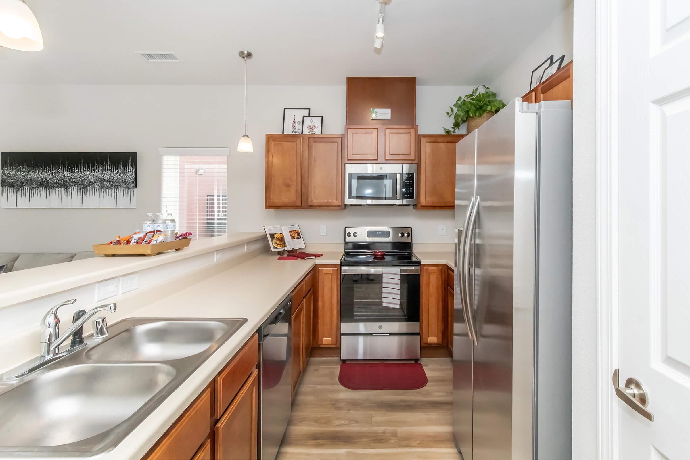 A modern kitchen featuring wooden cabinets, stainless steel appliances including an oven and refrigerator, a double sink, and a light-colored countertop. The kitchen includes a red mat on the floor and a decorative plant, with a view of a living area in the background.