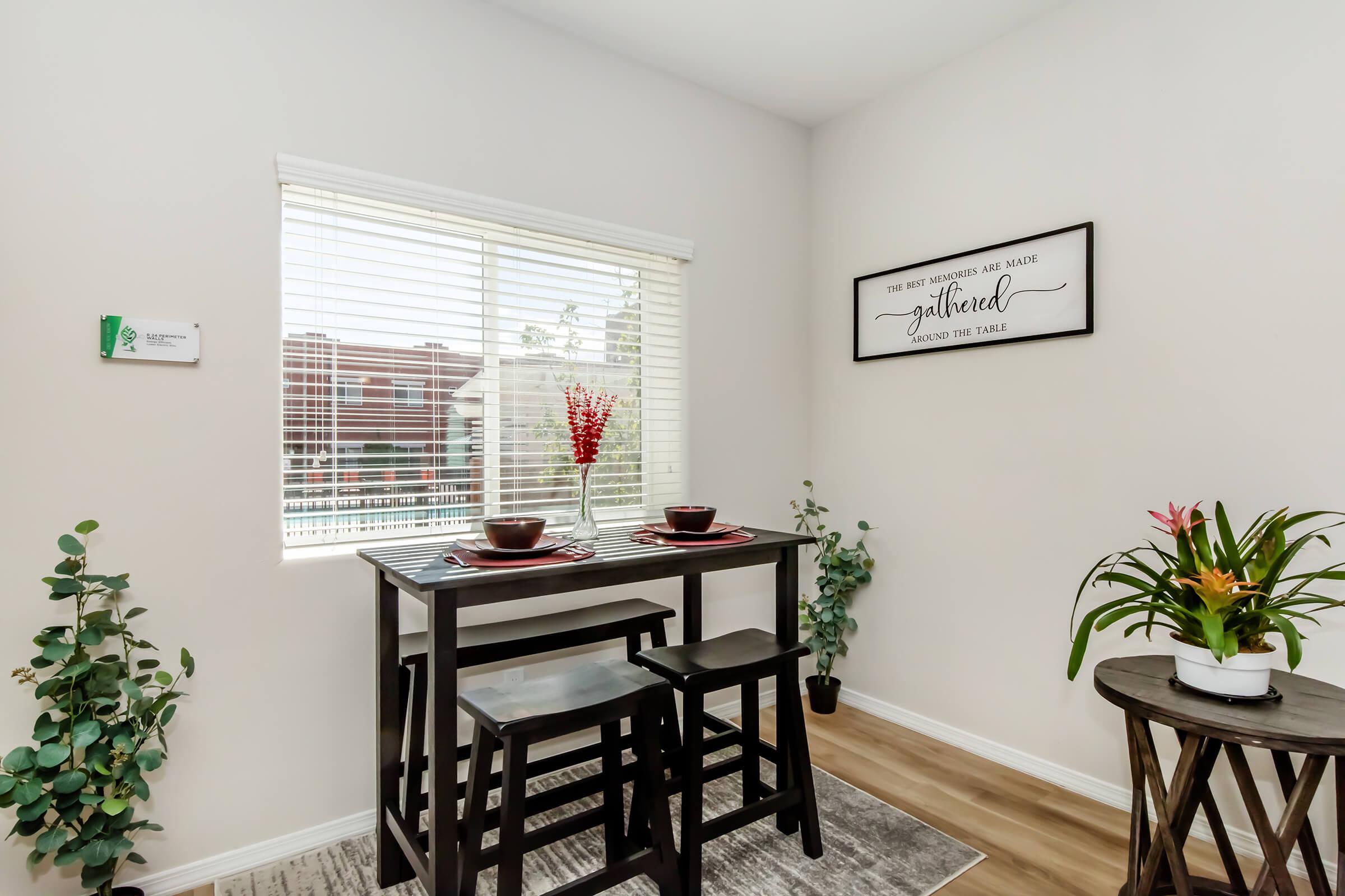 A small dining area with a black table and two stools, set with two plates and bowls. There's a window with white blinds, allowing natural light in. A framed decorative sign with the words "Gathered around the table" hangs on the wall. Two green plants are on either side of the table, enhancing the cozy atmosphere.