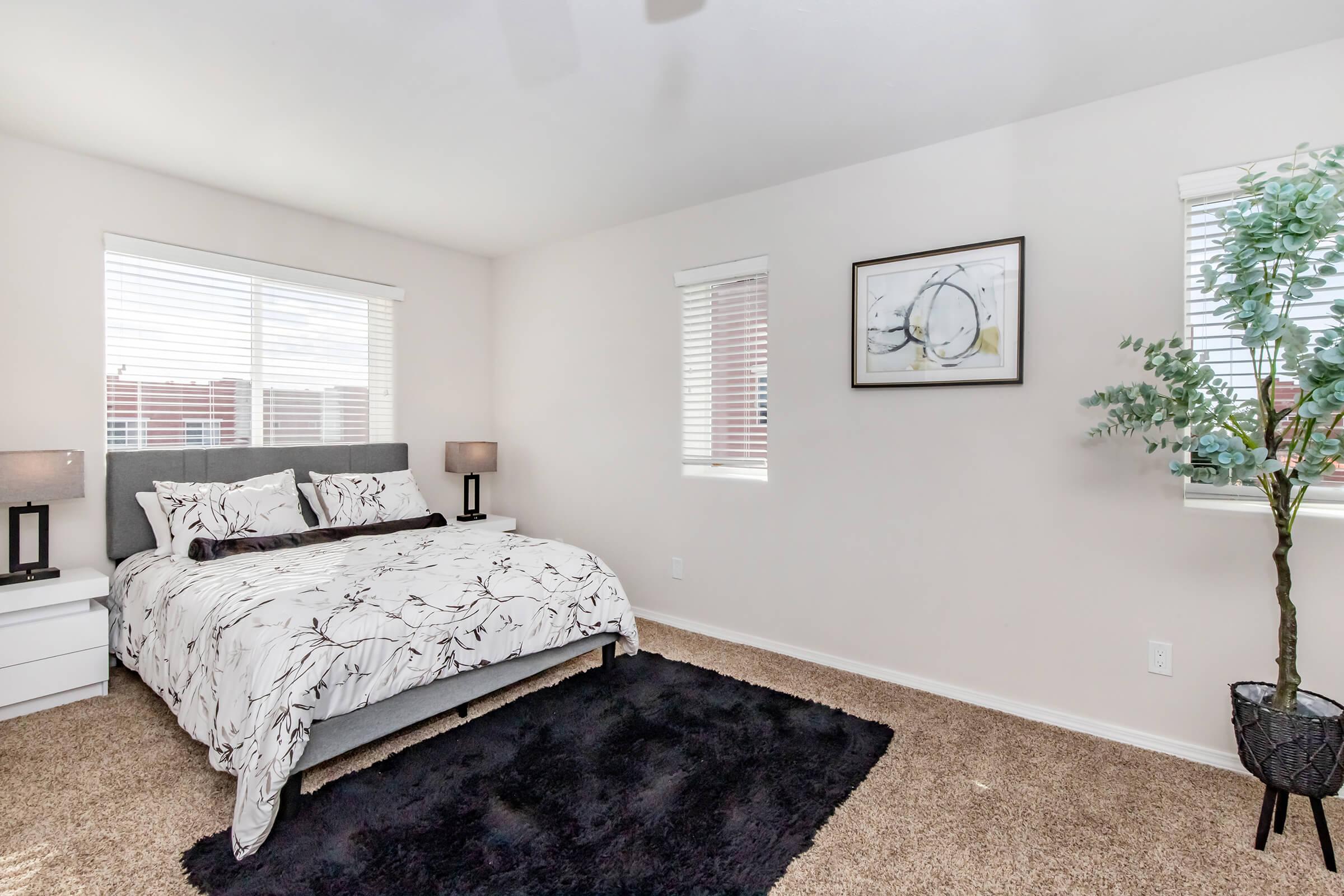 A cozy bedroom featuring a queen-sized bed with a floral patterned duvet cover, two bedside lamps, and a large black area rug. Natural light fills the room through two windows with white blinds, and a framed artwork hangs on the wall. A potted plant adds a touch of greenery.