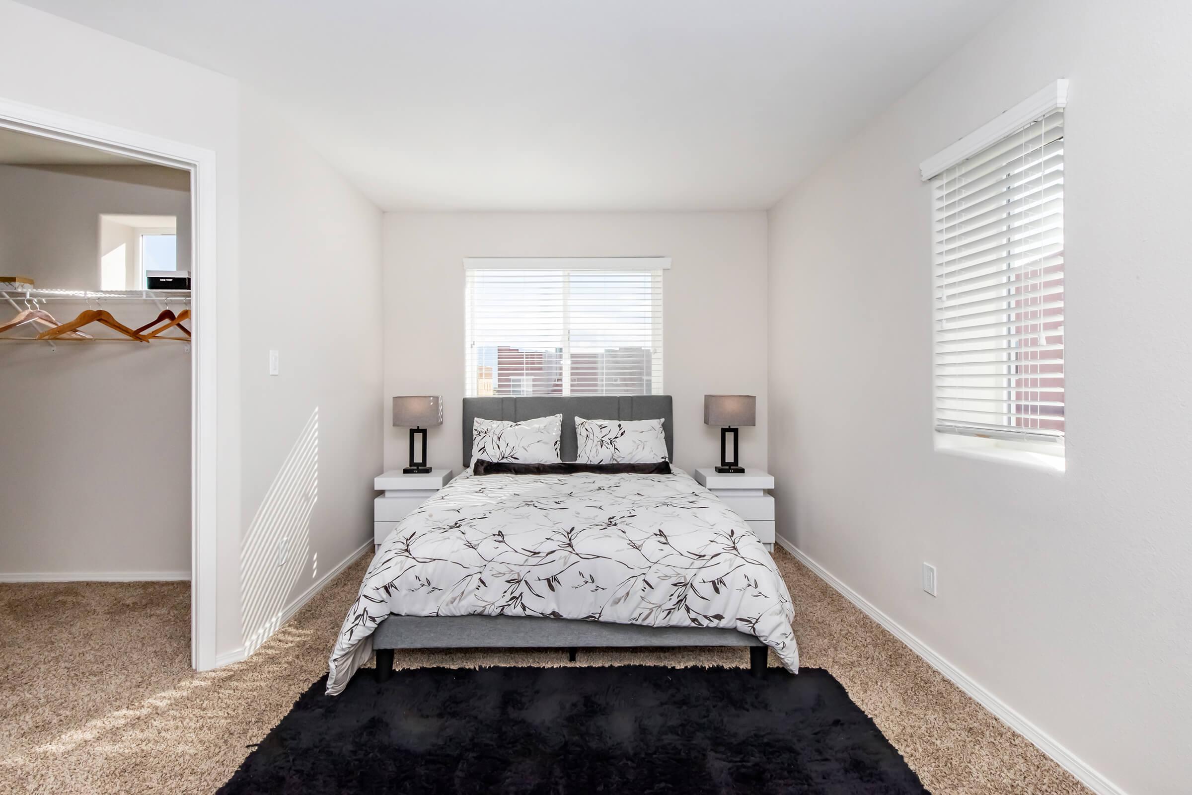 Bright and cozy bedroom featuring a double bed with a floral-patterned duvet, two bedside lamps, and large windows allowing natural light. The room is carpeted and has a closet with wooden hangers visible in the adjacent room. Neutral wall colors create a calm atmosphere.