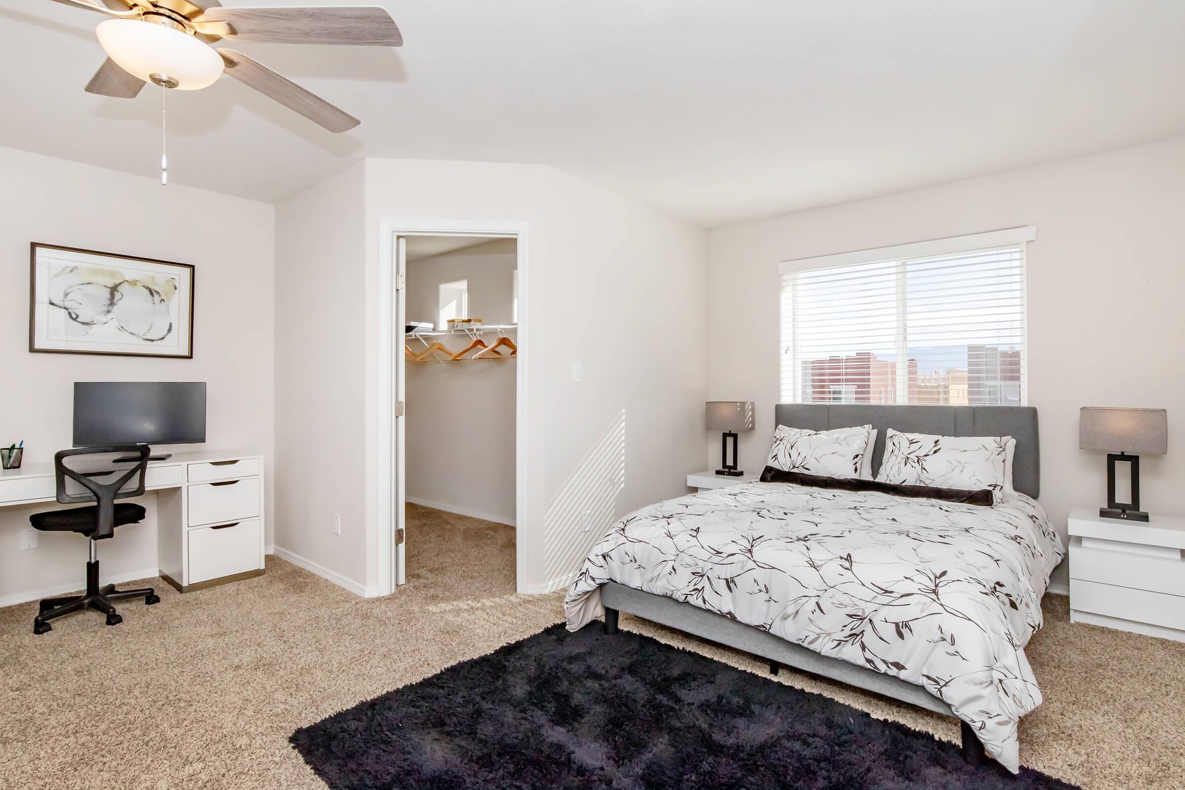 A cozy bedroom featuring a comfortable bed with floral bedding, a black rug, a desk with a chair, and a ceiling fan. There is a closet with hangers visible through an open door, and large windows letting in natural light. The walls are painted in neutral colors, creating a calm atmosphere.