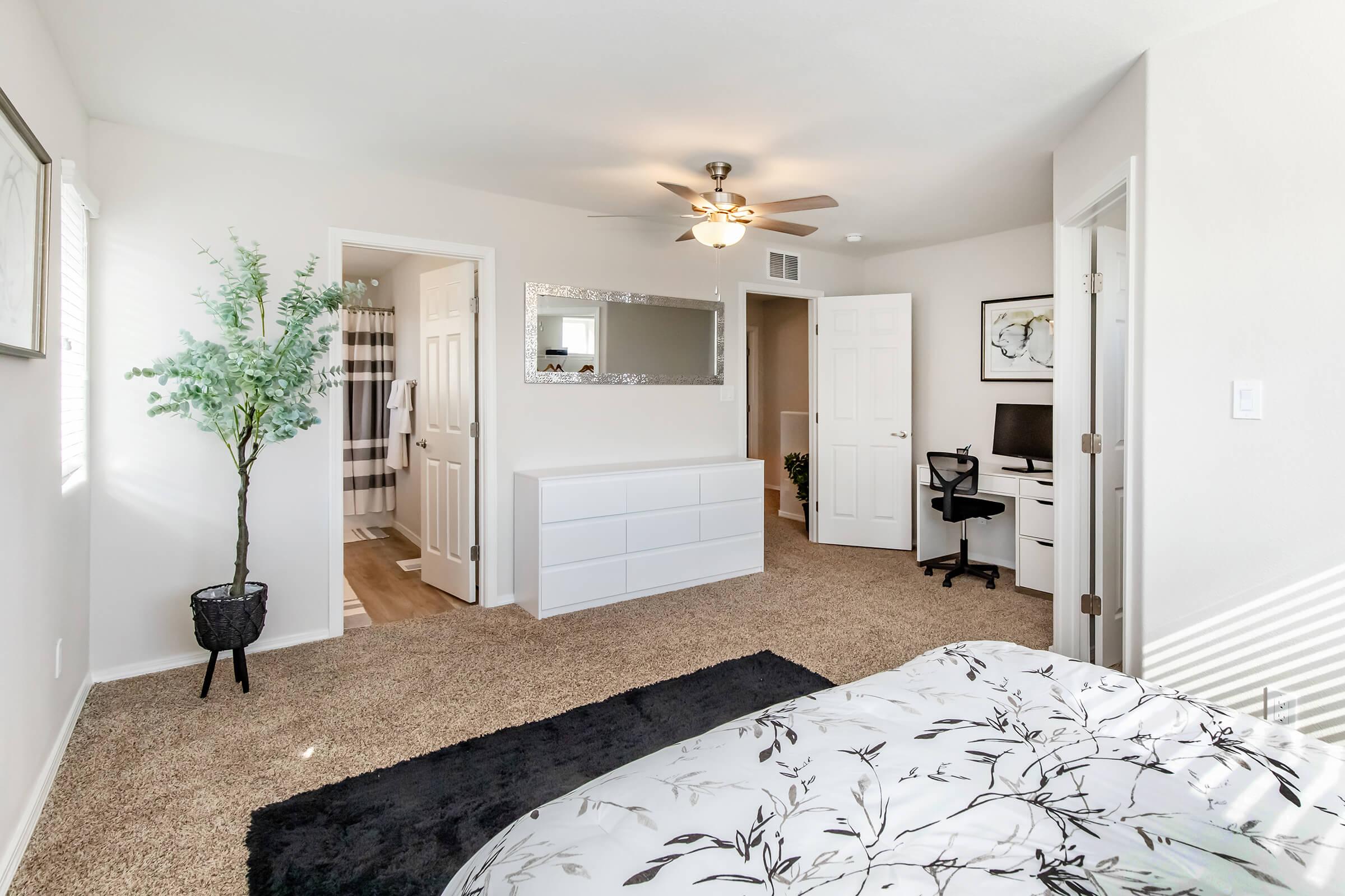 A spacious, well-lit bedroom featuring a large bed with a floral comforter, a white dresser, and a modern desk with a black chair. A potted plant adds a touch of greenery, while a bathroom door and a mirrored wall decorate the space. The room has light-colored walls and plush carpet flooring.