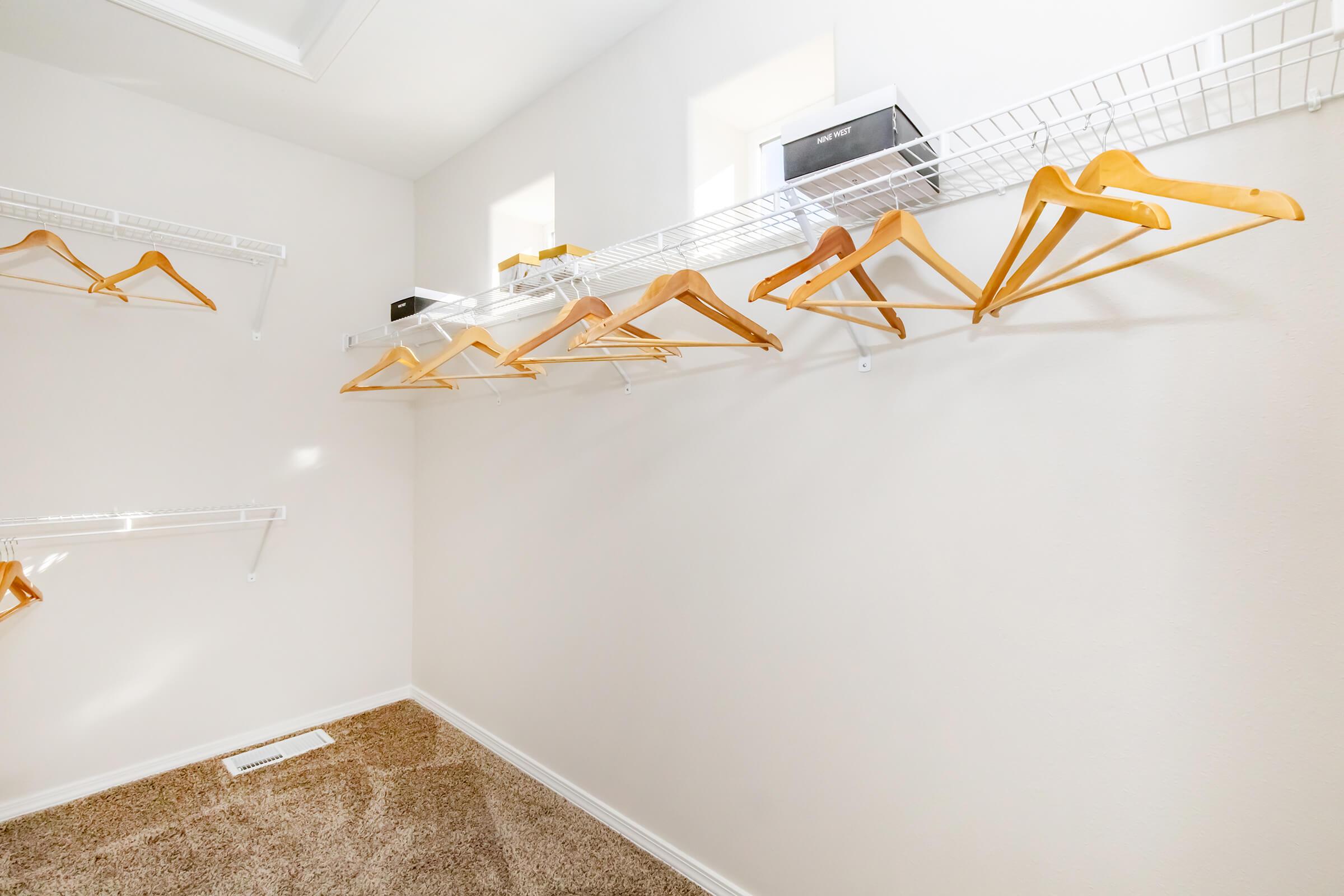 Empty closet with a beige carpet and white walls. Several wooden hangers are hanging on wire shelves along the wall, while a couple of small boxes are placed on the top shelf. Natural light comes in through small windows.