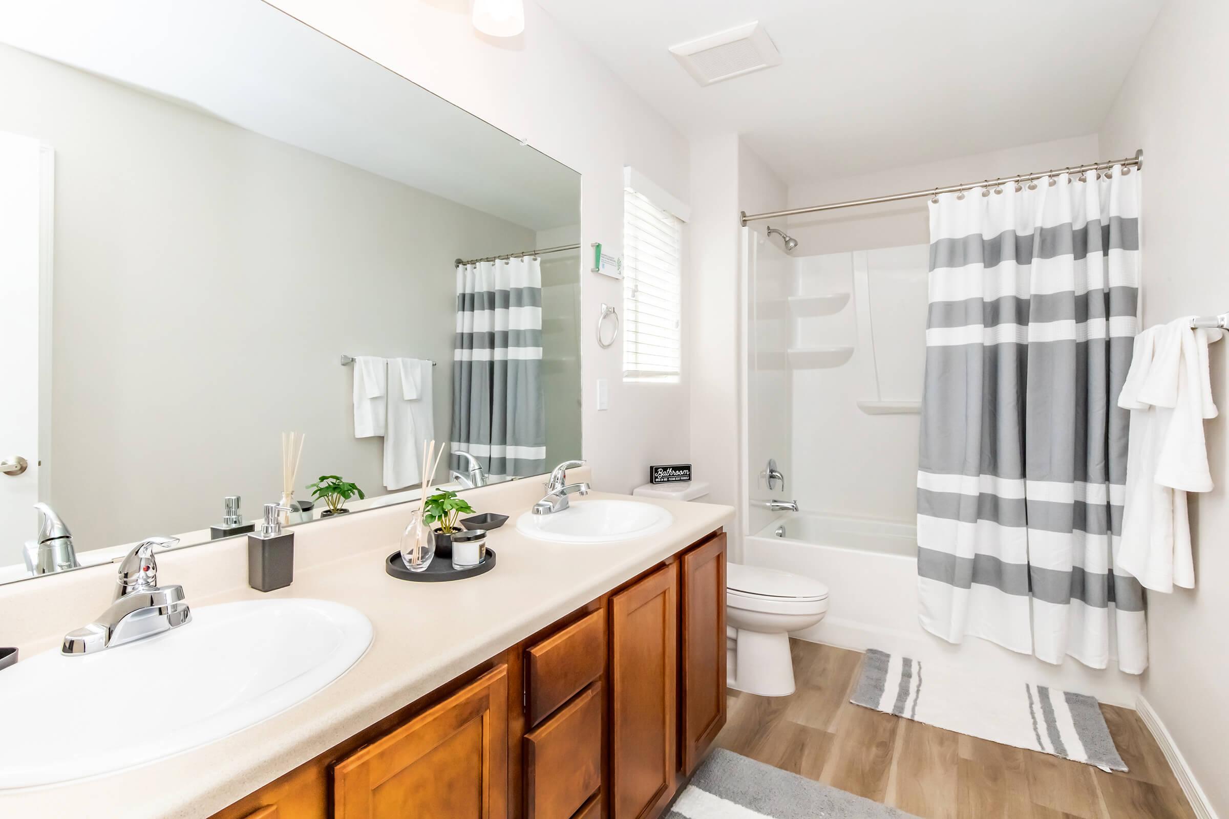 A modern bathroom featuring dual sinks with silver faucets, a large mirror, and light gray walls. The space includes a bathtub with a shower curtain sporting gray and white stripes, a towel rack, and plants arranged decoratively on the counter. Soft rugs are placed on the floor for comfort.