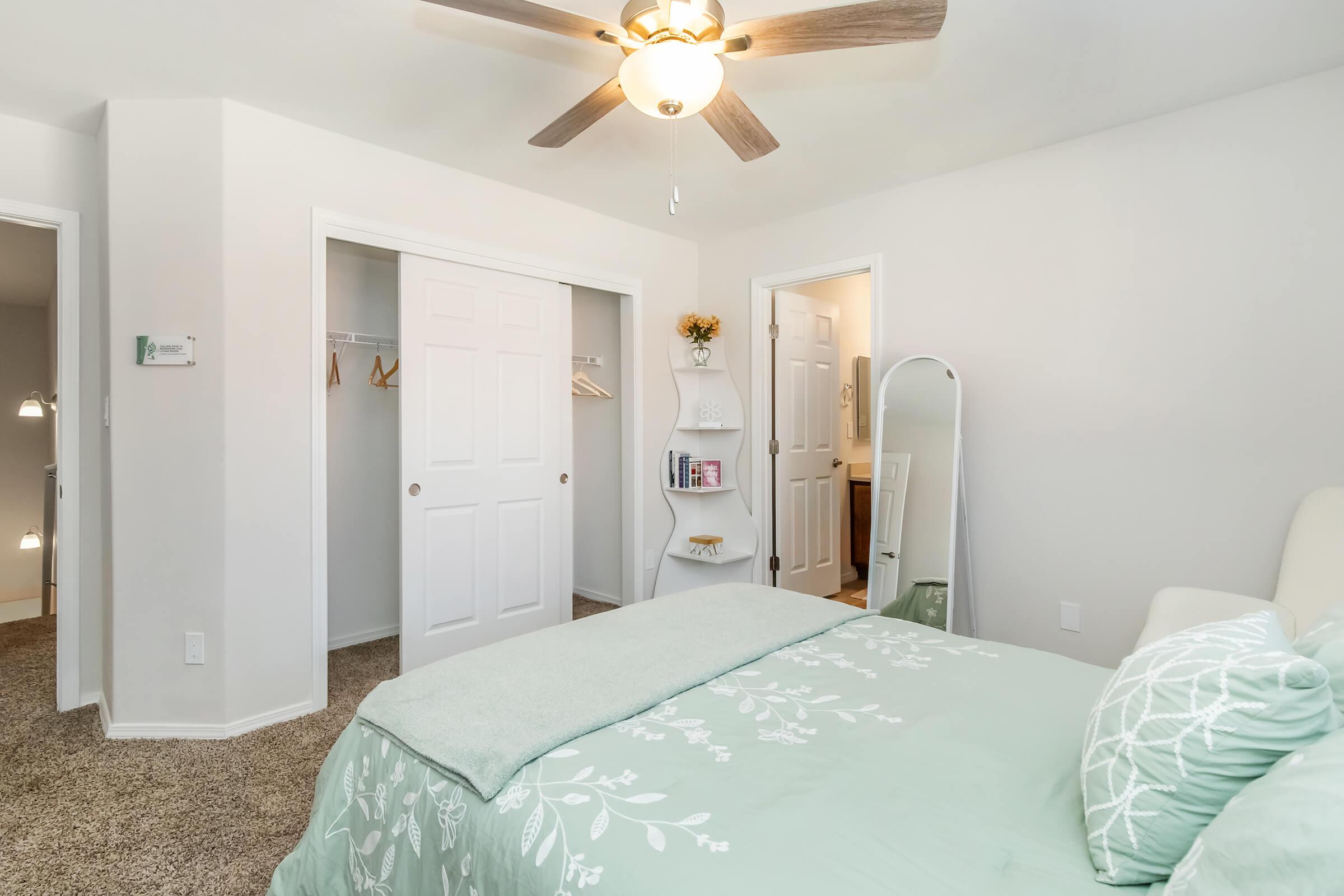 A cozy bedroom featuring a light green bedspread and a decorative white pillow. The room has a ceiling fan, a full-length mirror, and white closet doors. A shelving unit displays books and decor, while two doorways lead to adjacent spaces. Soft, neutral tones create a welcoming atmosphere.