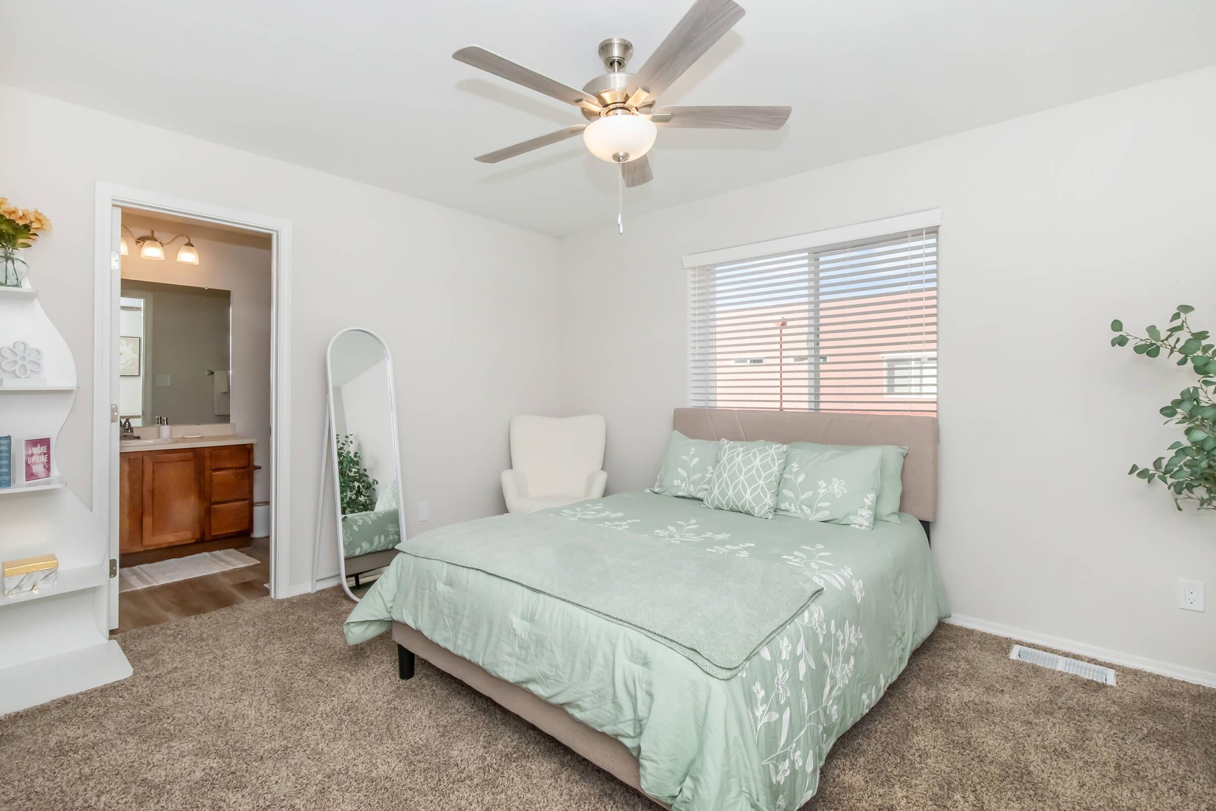 A cozy bedroom featuring a large bed with a light green cover, a ceiling fan above, and a window with blinds. There's a full-length mirror and a shelving unit next to the bed. The room has soft carpet flooring and a bathroom visible through an open door, creating a welcoming atmosphere.