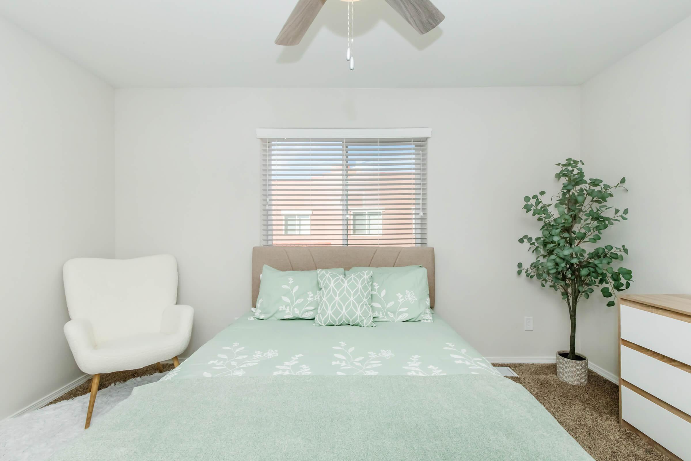 A cozy bedroom featuring a bed with light green bedding, decorative pillows, and a white armchair. A potted plant and a dresser are present, with a window covered by blinds providing natural light. The walls are painted in a neutral color, creating a calm and inviting atmosphere.