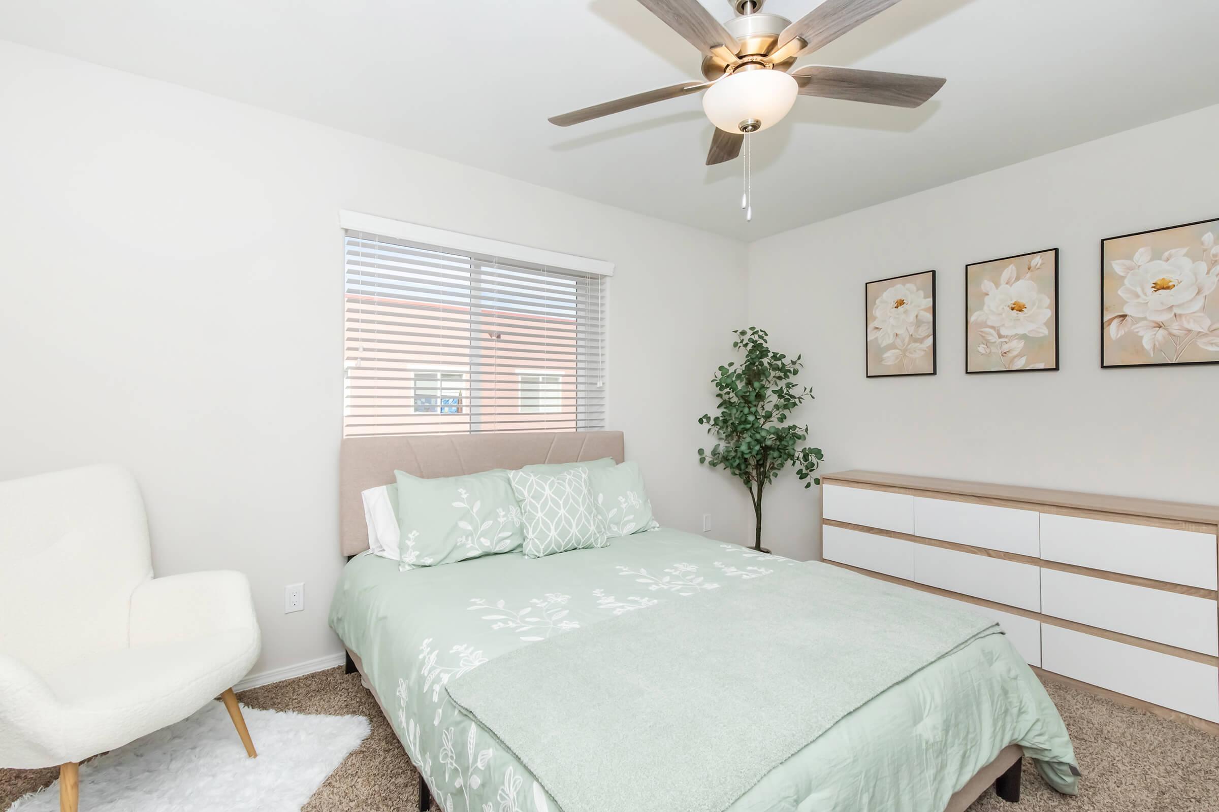 A cozy bedroom featuring a light green bedspread on a bed with decorative pillows. The walls are adorned with three floral art pieces. A window with blinds allows natural light, and a small indoor plant adds a touch of greenery. A white armchair and a sleek dresser complete the minimalist decor.