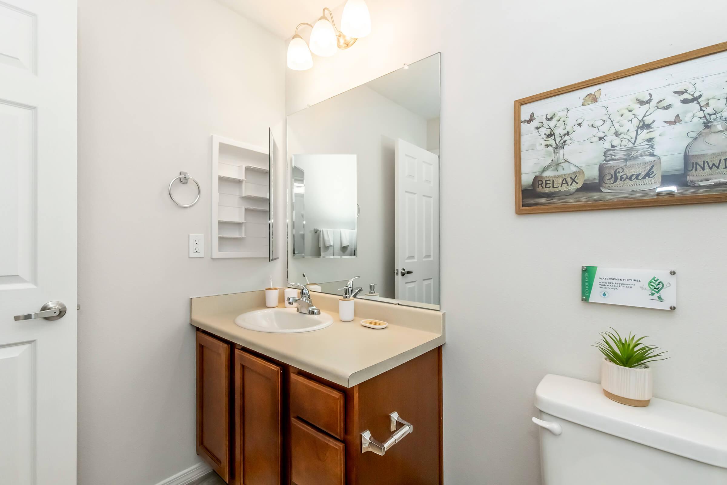 A modern bathroom with a light-colored vanity, a large mirror above the sink, and wooden cabinetry. There is a decorative wall art with the words "RELAX" and "SOAK," a plant on the countertop, and a towel rack beside the sink. The scene is well-lit and displays a clean, inviting atmosphere.