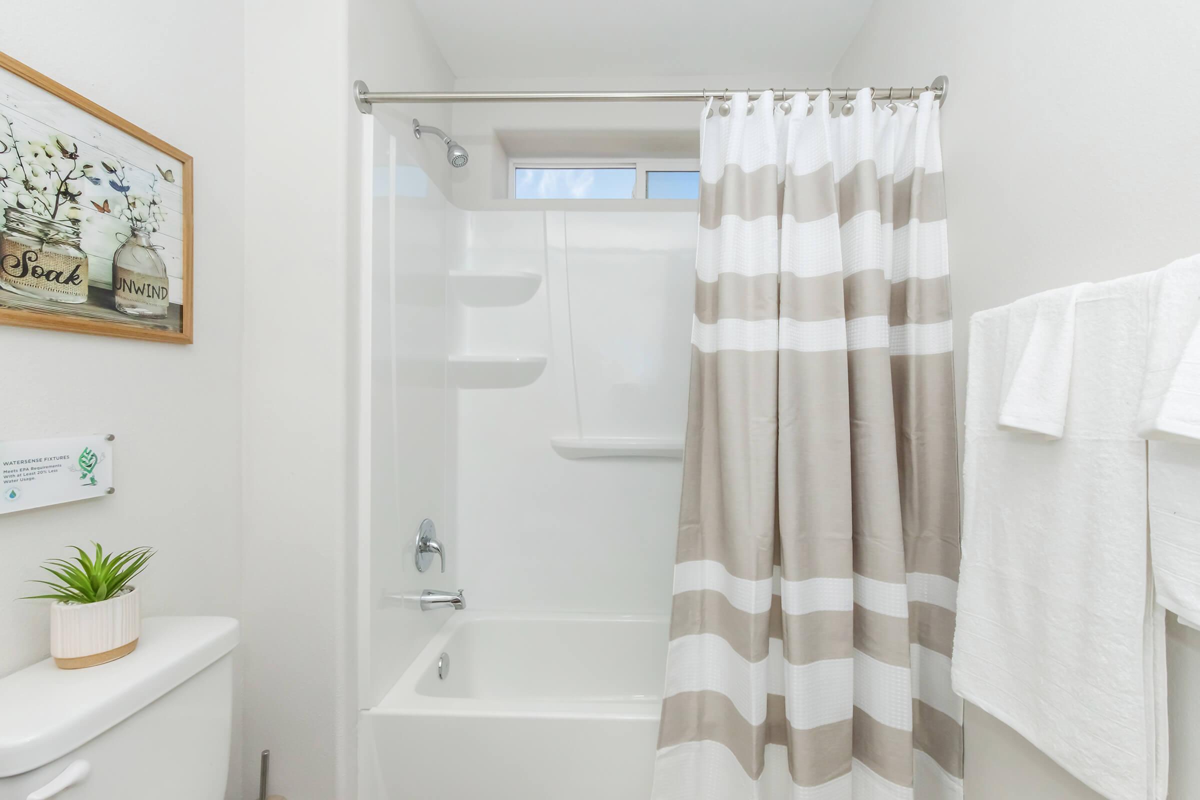 A modern bathroom featuring a bathtub with a shower curtain in beige and white stripes. There's a small potted plant on the toilet tank, a framed floral print on the wall, and neatly arranged white towels hanging on a rod, creating a clean and welcoming atmosphere.
