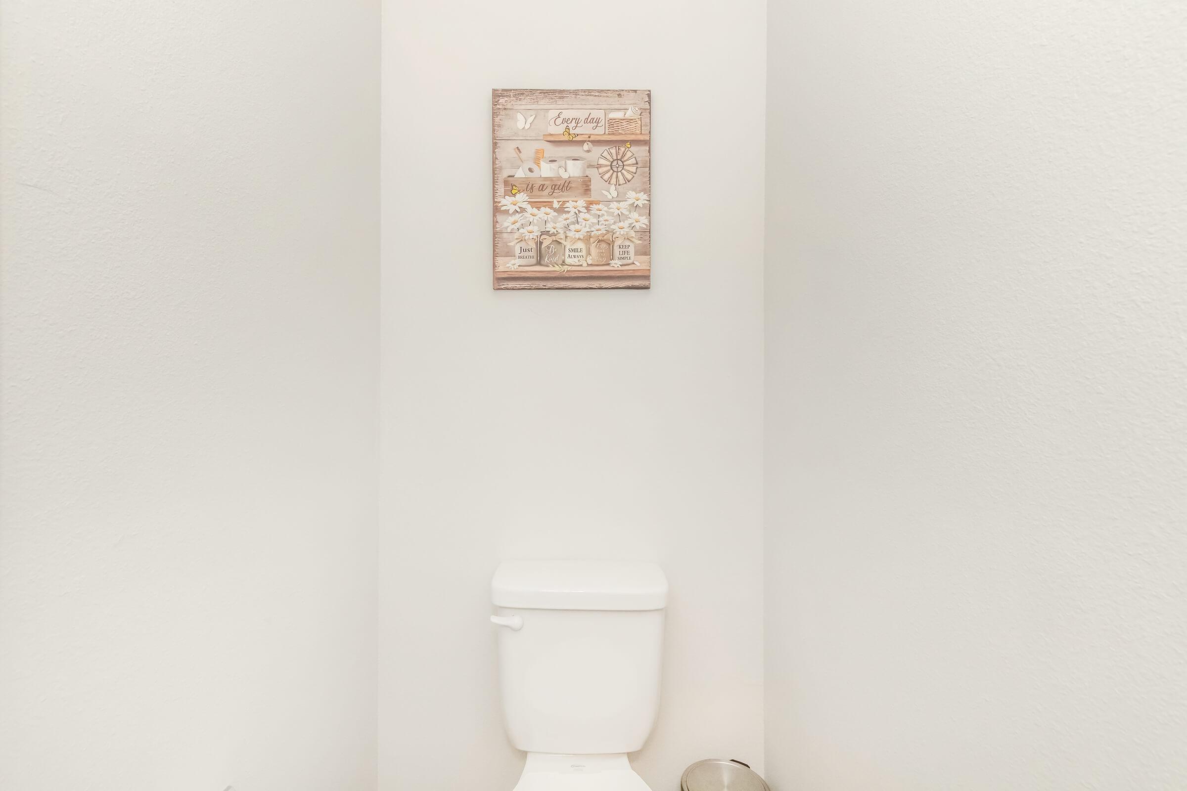 A clean, minimalistic bathroom setting featuring a white toilet against a plain white wall. Above the toilet is a framed decorative sign featuring floral imagery and text. A small metal trash bin is placed on the floor nearby, maintaining the tidy and simple aesthetic of the space.