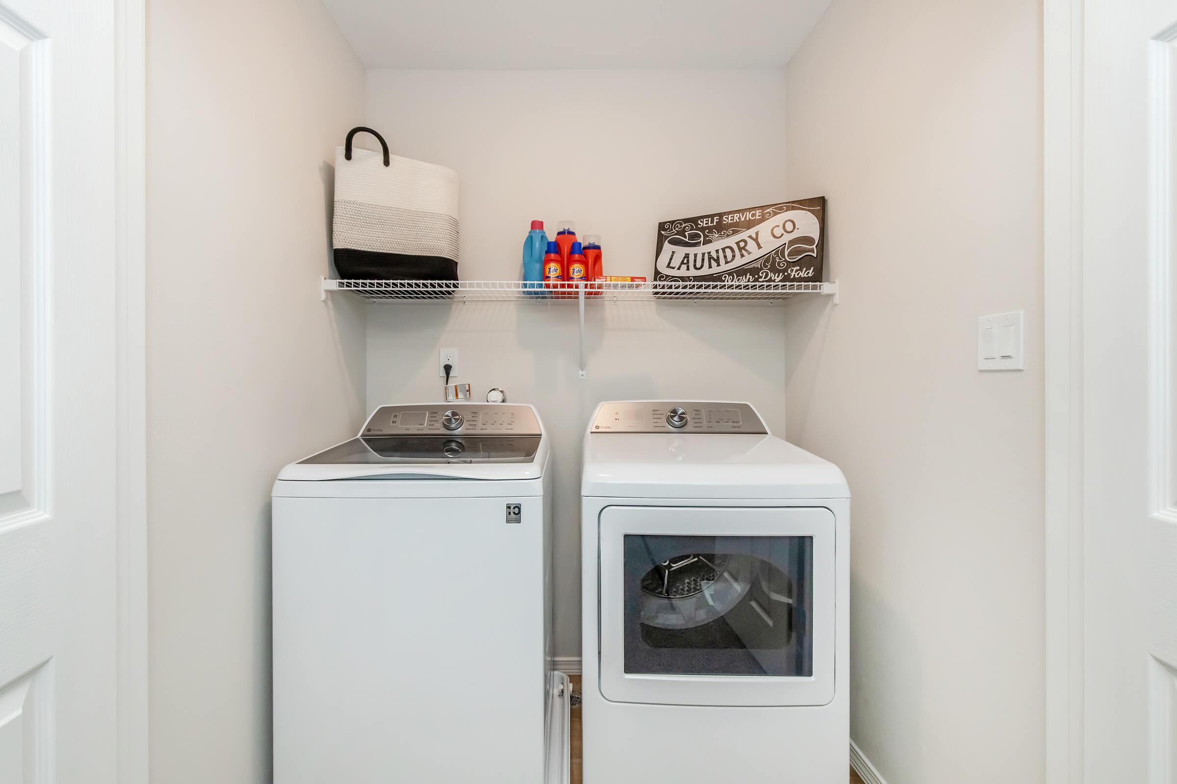 A neat laundry room featuring a washing machine and dryer side by side. Above them, there is a shelf with laundry supplies, including detergents. A stylish laundry sign is mounted on the wall. The walls are painted a light color, creating a bright and clean atmosphere.