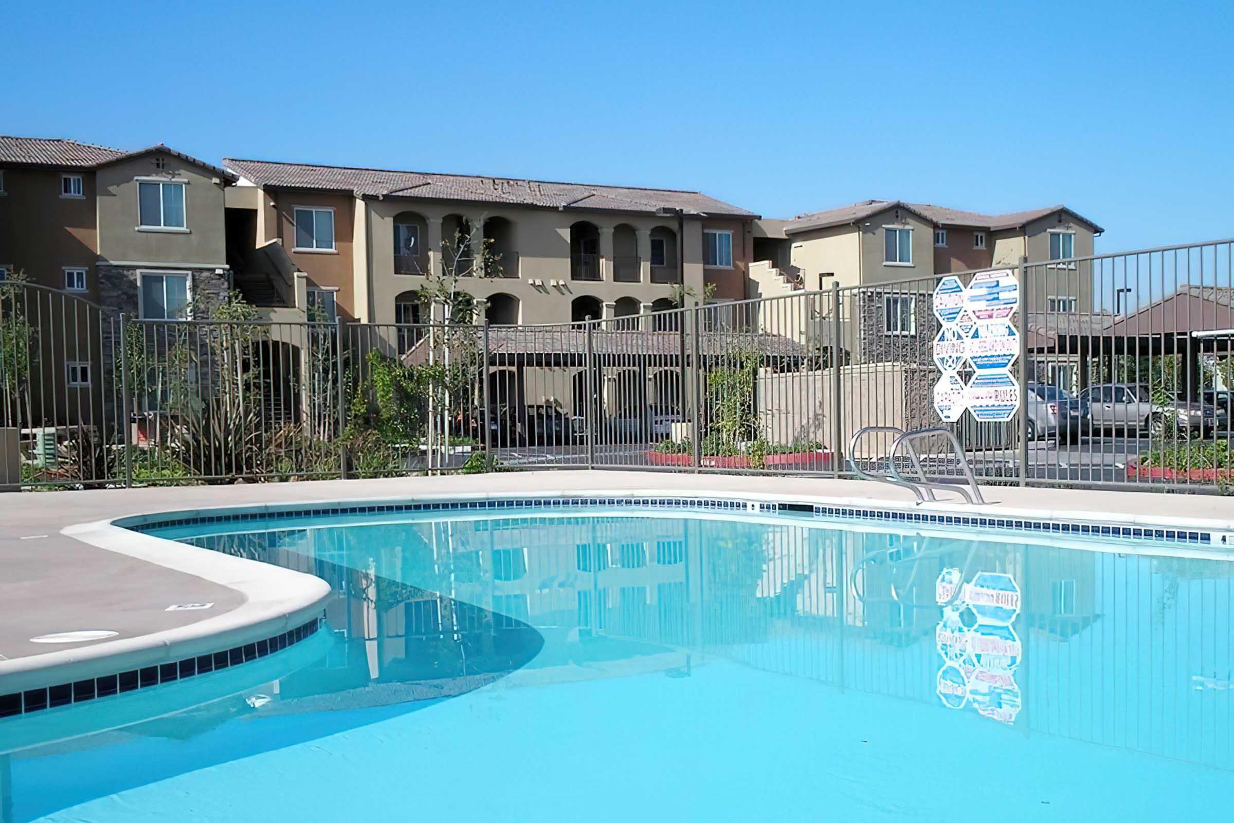 A clear blue swimming pool with a surrounding fence, positioned in front of a residential building with multiple apartments.
