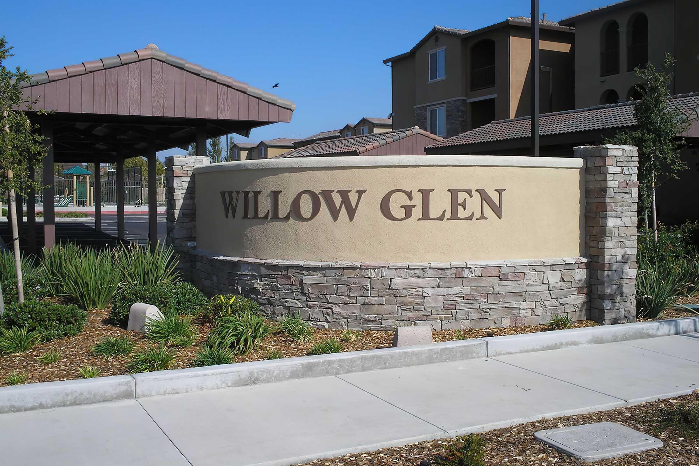 Entrance sign for Willow Glen, featuring stone and stucco design, surrounded by greenery and a covered structure.