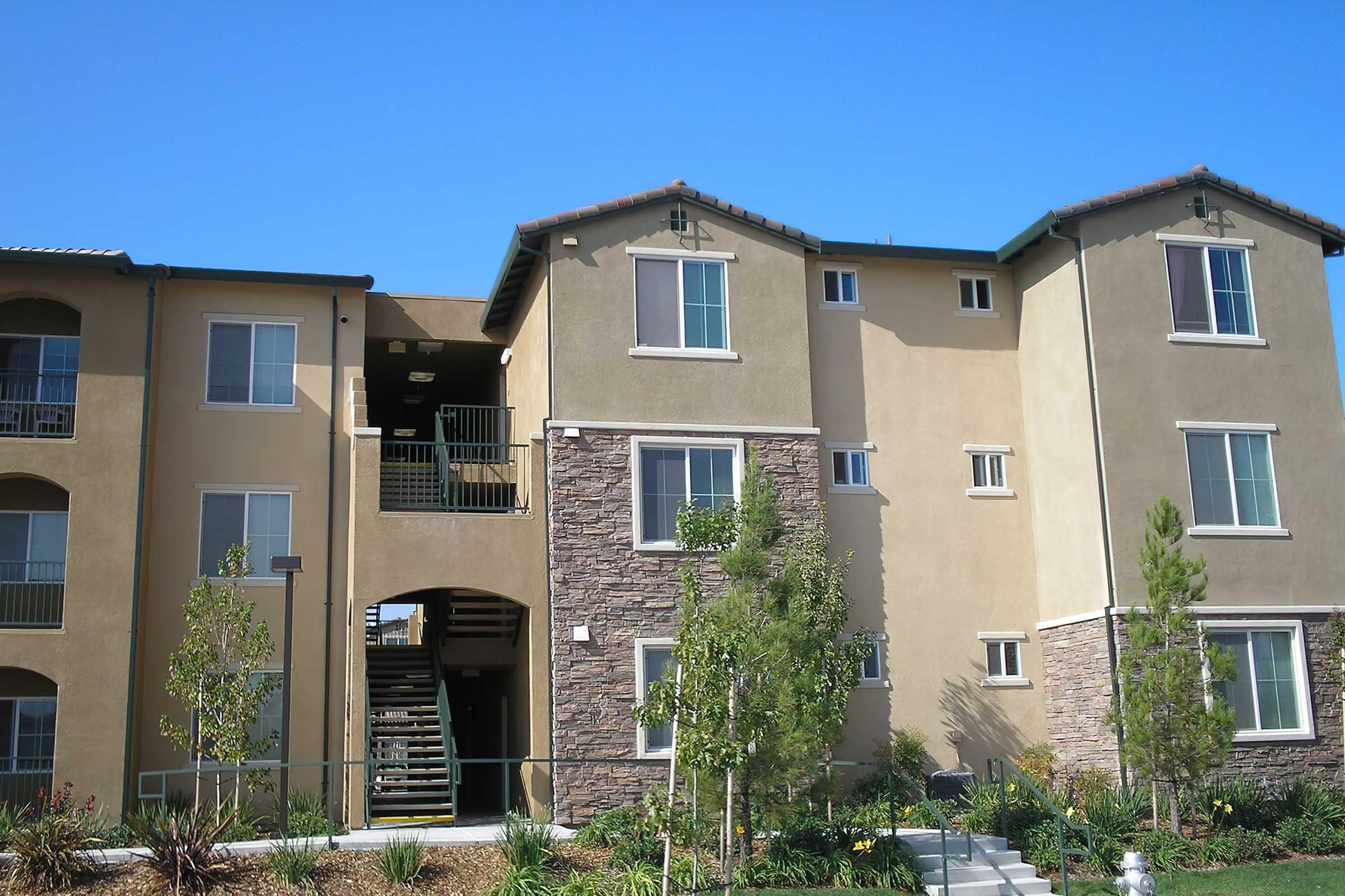 A modern residential building featuring a combination of tan stucco and stone facade. The structure has multiple levels, with balconies and large windows.