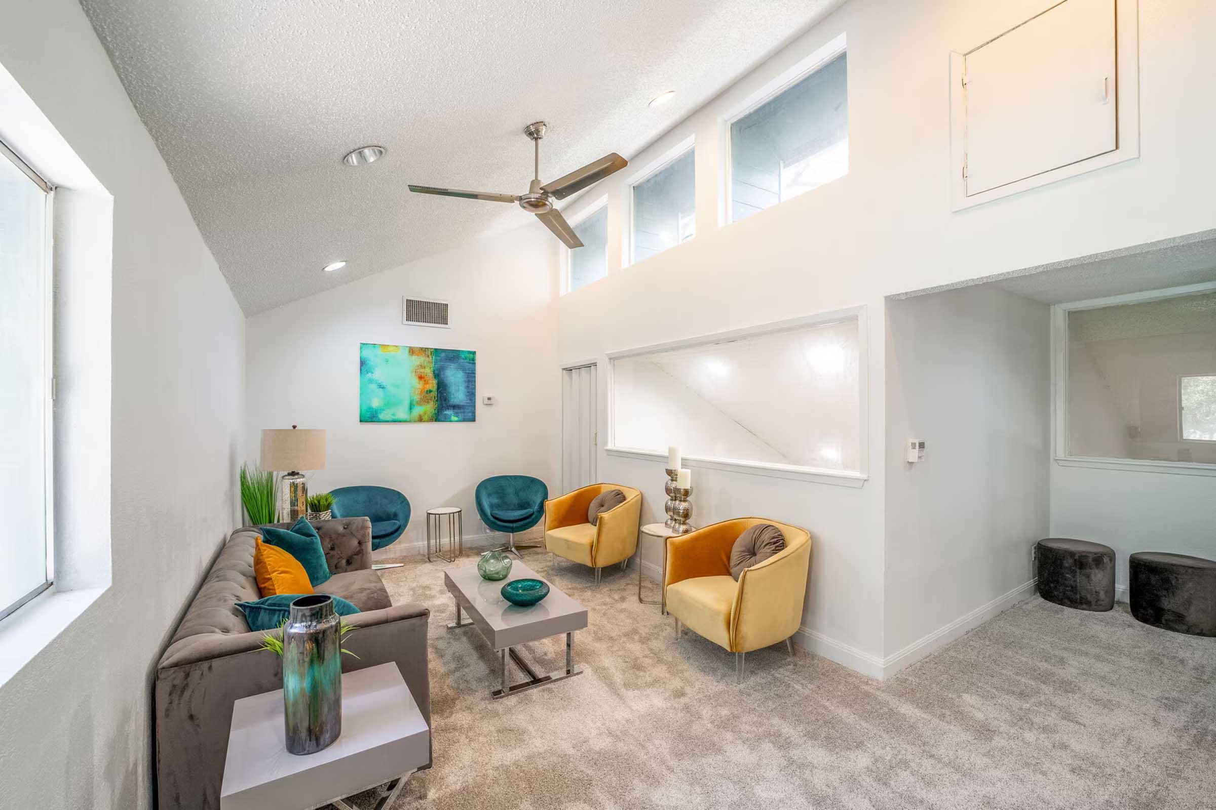 A modern and cozy living room with a grey sofa, two vibrant orange and blue chairs, and large windows allowing natural light. A ceiling fan hangs above, and colorful abstract artwork adorns the walls. The space features a light carpet and sleek furniture, creating a welcoming atmosphere.
