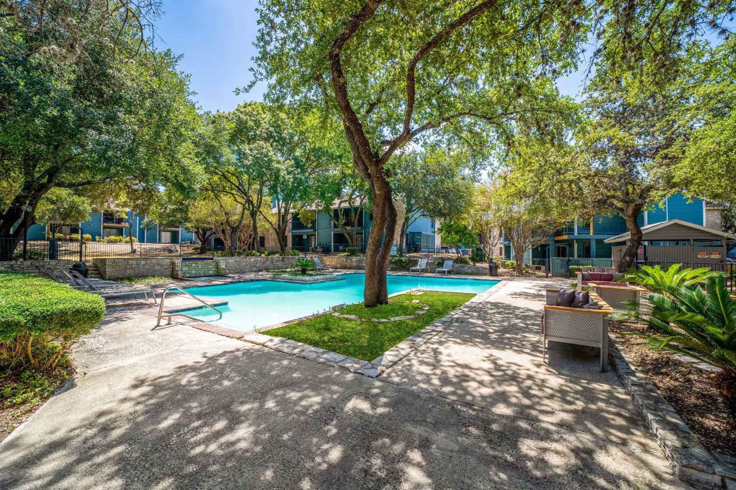 A serene outdoor pool area surrounded by lush green trees and landscaping. Comfortable seating areas are positioned nearby, creating a relaxing atmosphere. The pool is inviting, with clear blue water reflecting the sunlight, while the surrounding apartment buildings add to the tranquil setting.