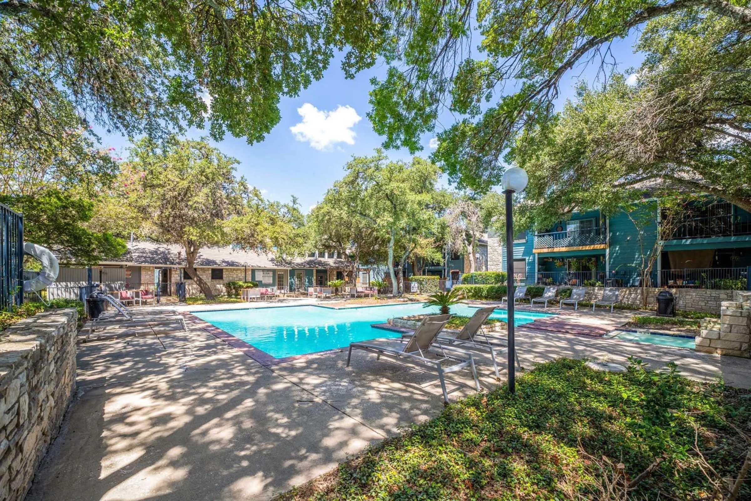 A scenic view of a swimming pool surrounded by lounge chairs and lush greenery. Trees provide shade, and the area features stone walls and nearby buildings. The sky is clear with a few clouds, creating a relaxing atmosphere for leisure and enjoyment.