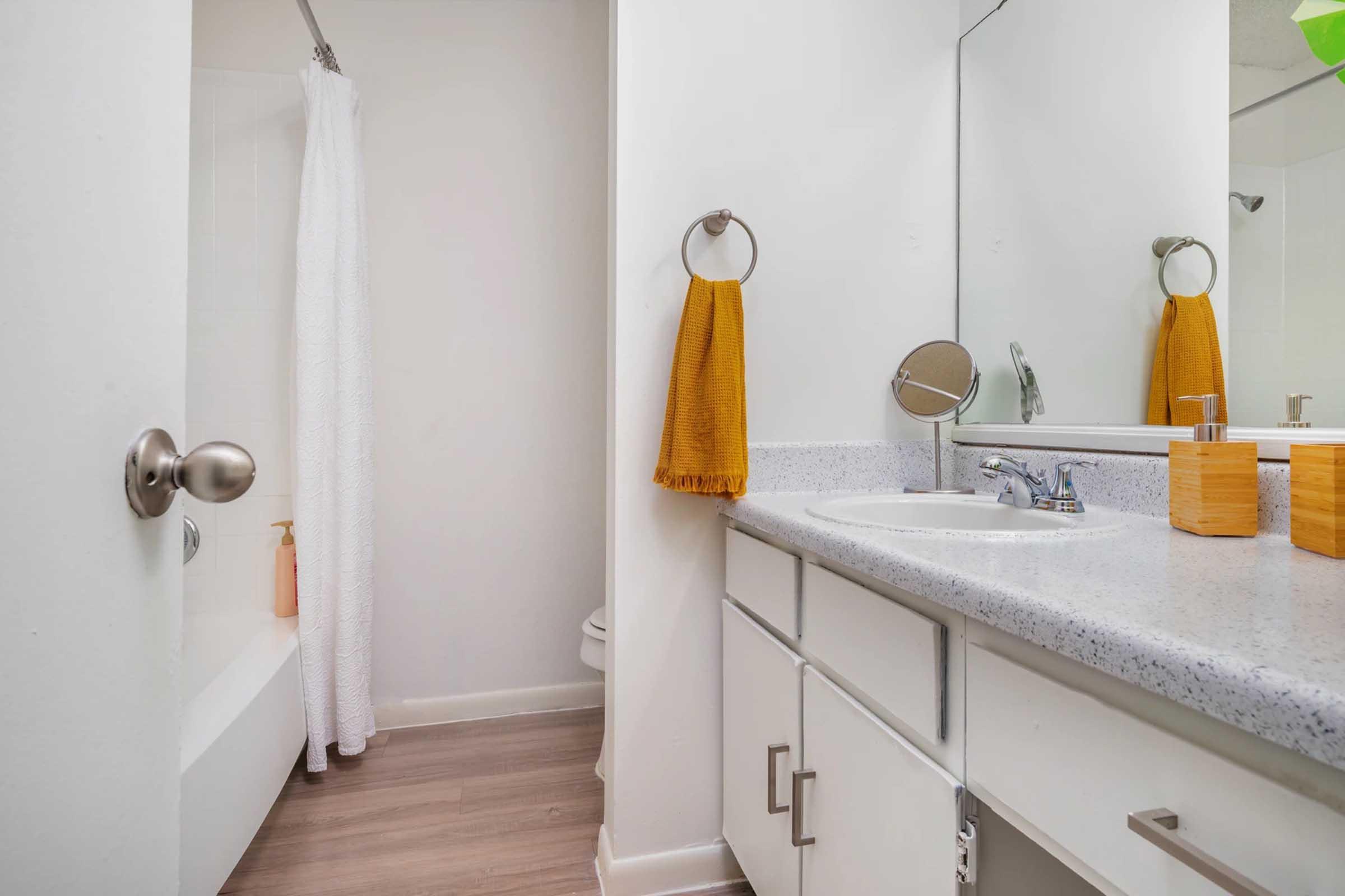 A modern bathroom featuring a bathtub with a white shower curtain, a sink with a speckled countertop, a mirror, a towel rack with an orange towel, and wood storage boxes. The decor is minimalistic with light colors and wooden flooring.