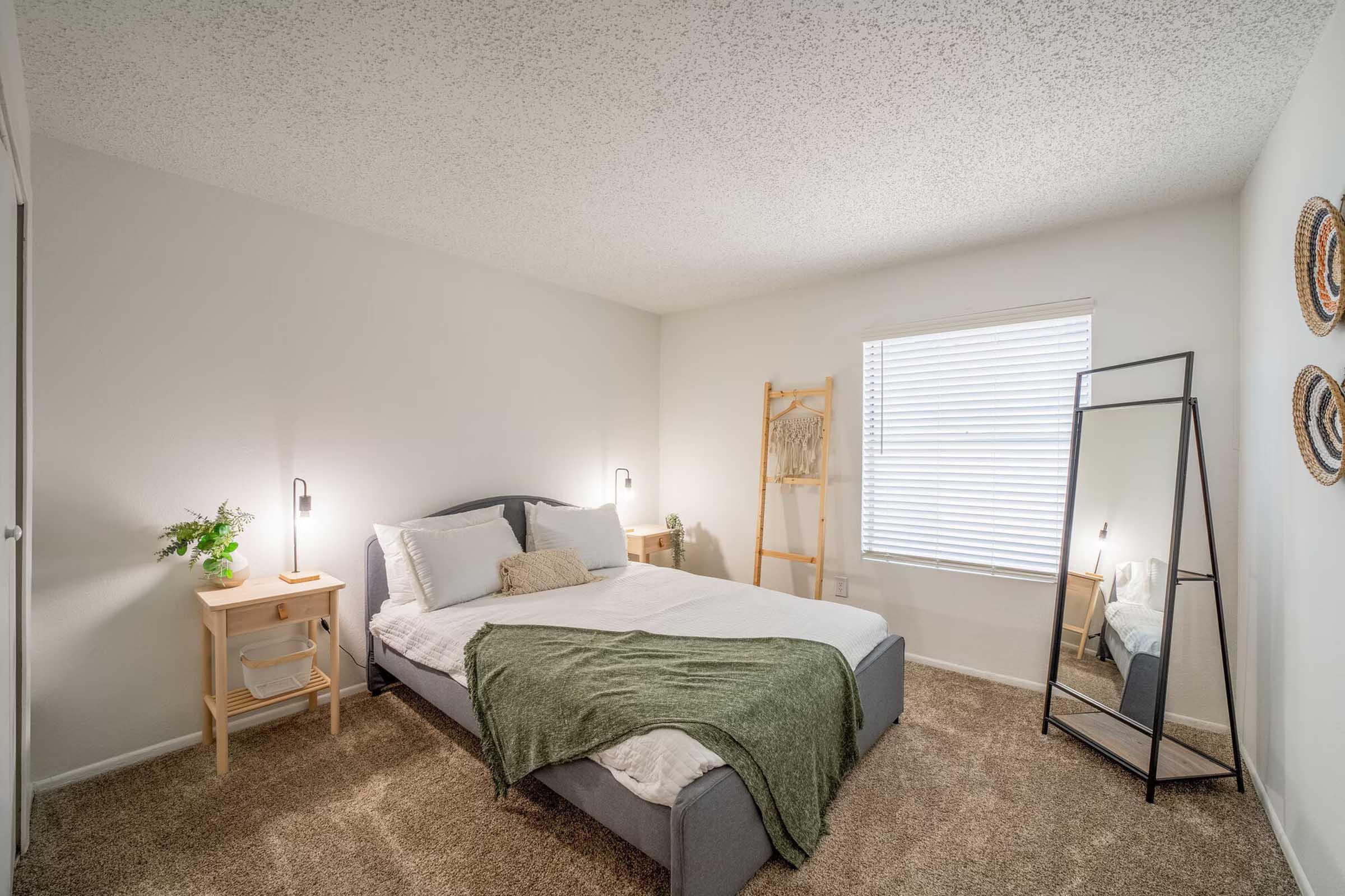 A cozy bedroom featuring a double bed with a green blanket, a small wooden nightstand with a plant, and a tall mirror. Soft, neutral walls and a textured ceiling complement the room's serene atmosphere. Natural light filters in through a window with blinds, enhancing the inviting space.