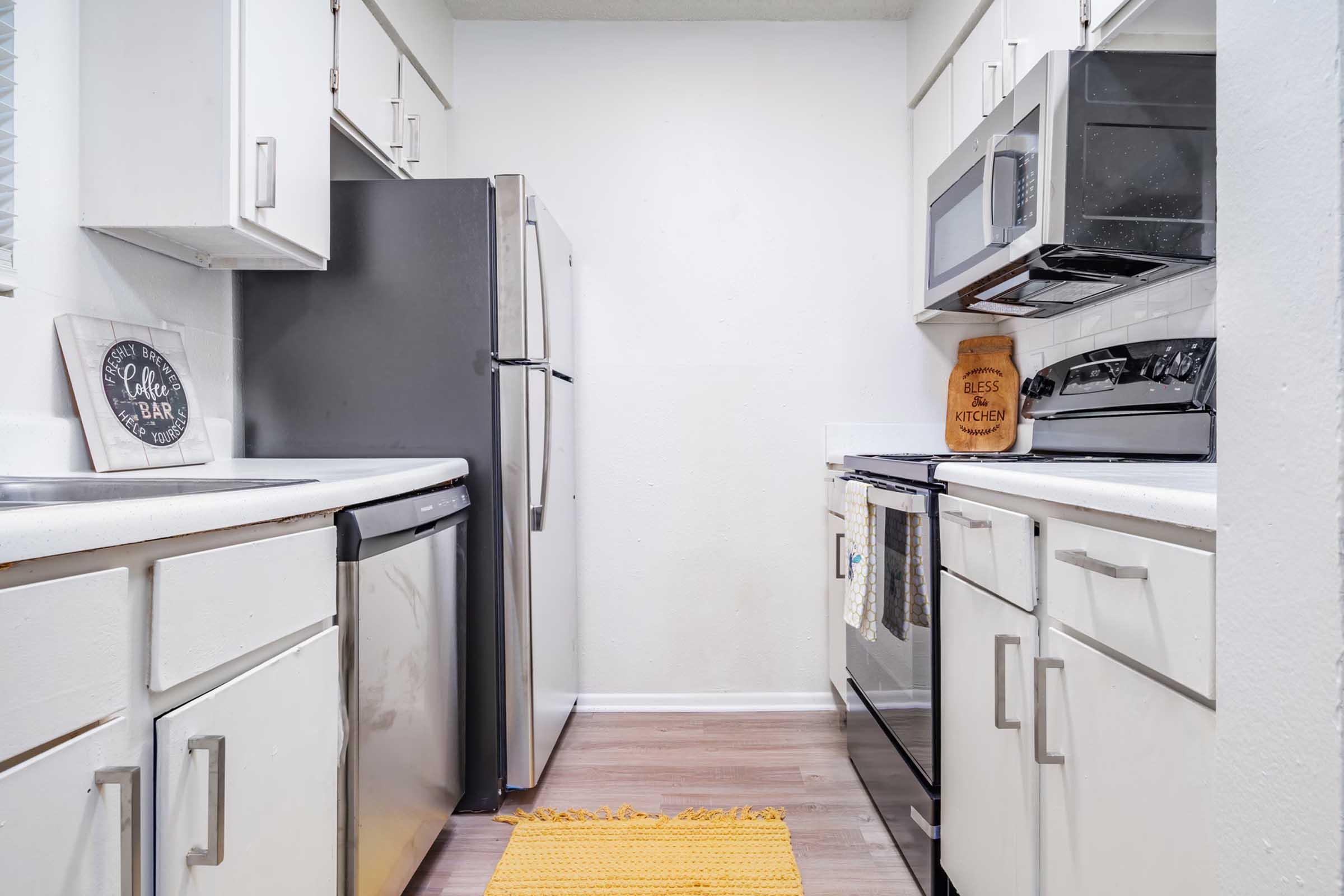 A clean, modern kitchen featuring white cabinets, stainless steel appliances, and a wooden floor. The space includes a refrigerator, stove, microwave, and sink, with a yellow rug on the floor. A decorative sign on the wall adds a welcoming touch.