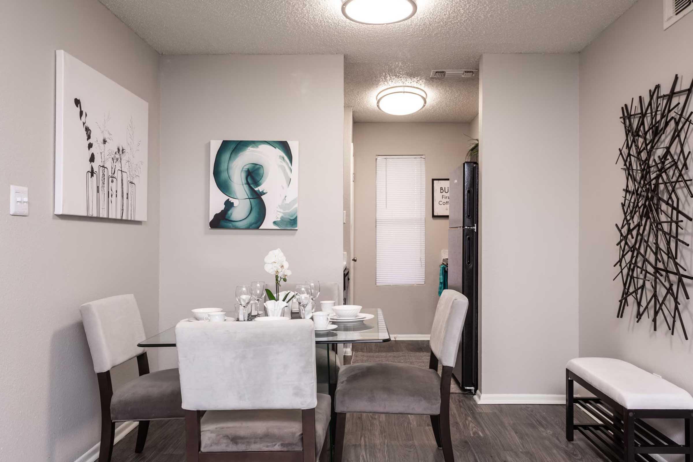 A modern dining area features a glass table set with dishes and silverware, surrounded by four chairs. On the walls, there are two elegant artworks—one with a cityscape design and another with a swirling teal pattern. A light fixture is visible above, along with a glimpse of a kitchen in the background.